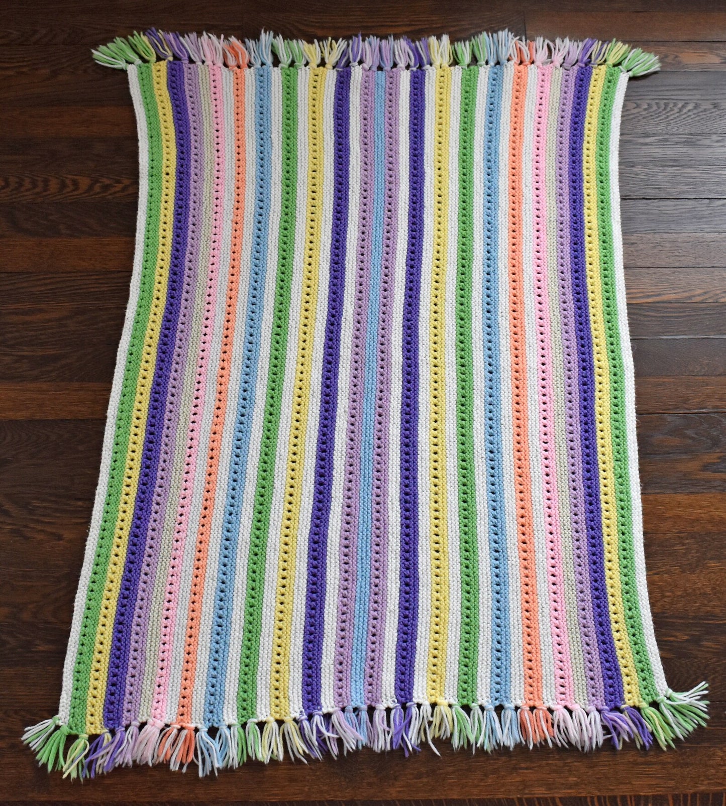 A white with dark and light purple, pink, blue, green, yellow, cream, and orange striped fringed blanket spread out on a wood floor.