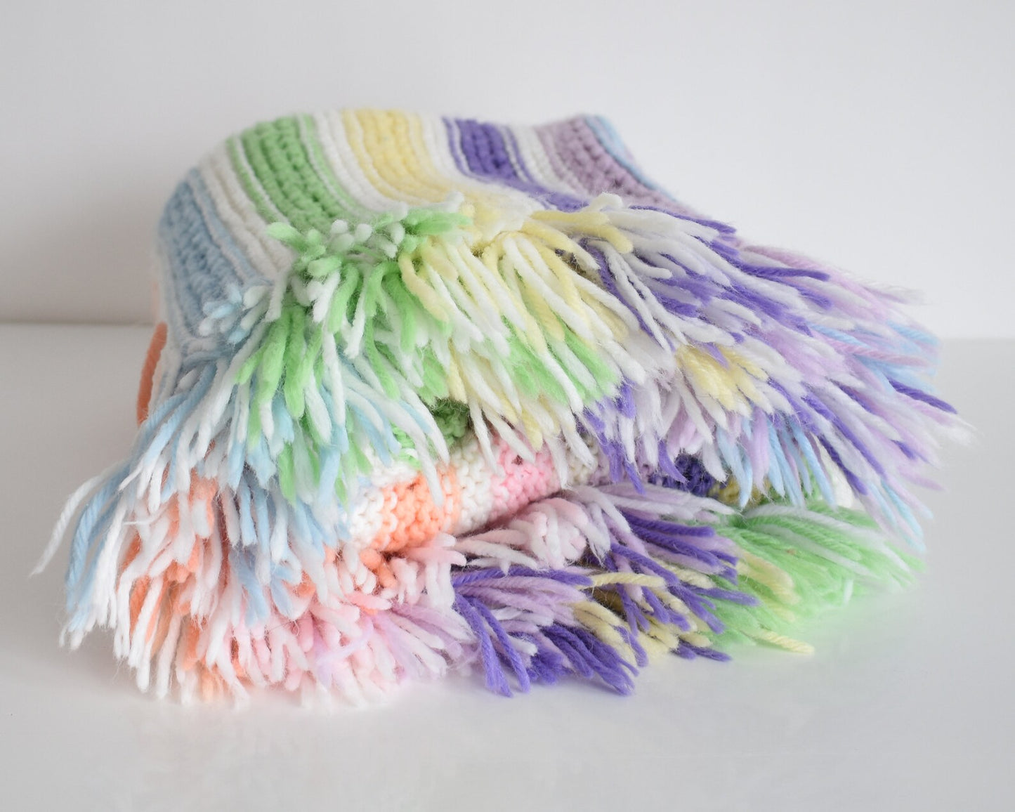 A white with dark and light purple, pink, blue, green, yellow, cream, and orange striped fringed blanket folded up on a white table.