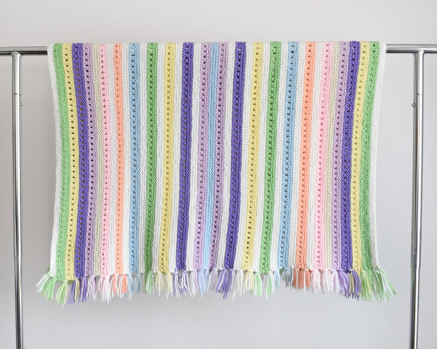 A white with dark and light purple, pink, blue, green, yellow, cream, and orange striped fringed blanket folded on a clothing rack