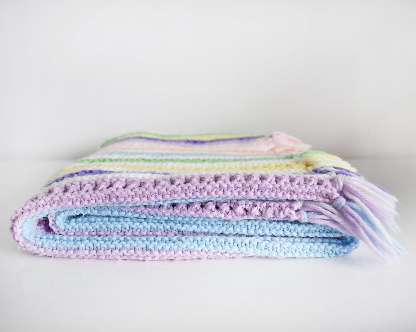 A white with dark and light purple, pink, blue, green, yellow, cream, and orange striped fringed blanket folded up on a white table.
