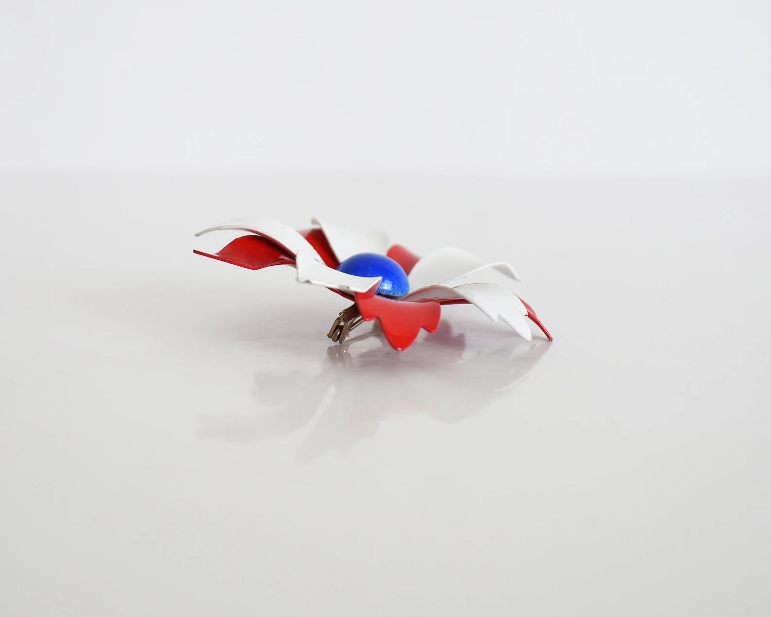 Side view of a vintage 60s mod floral brooch that has red and white petals with a blue rounded center. White and red backing with pin closure.
