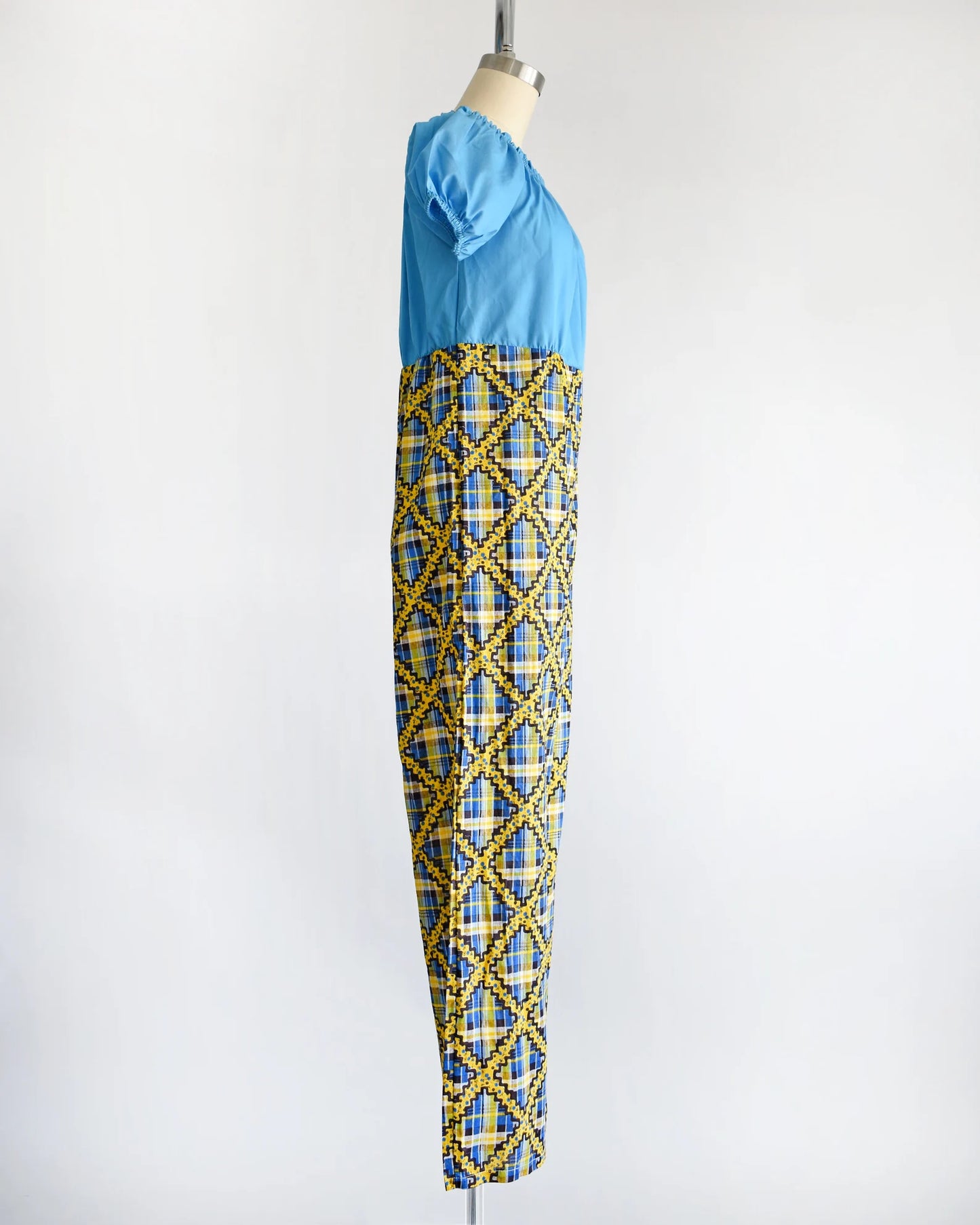 Side view of a vintage 70s jumpsuit that has a blue bodice with smocked elastic neckline and puff sleeves. The pants are a blue, black, and yellow plaid, with a ric-rac dotted yellow and black print on top of the plaid.