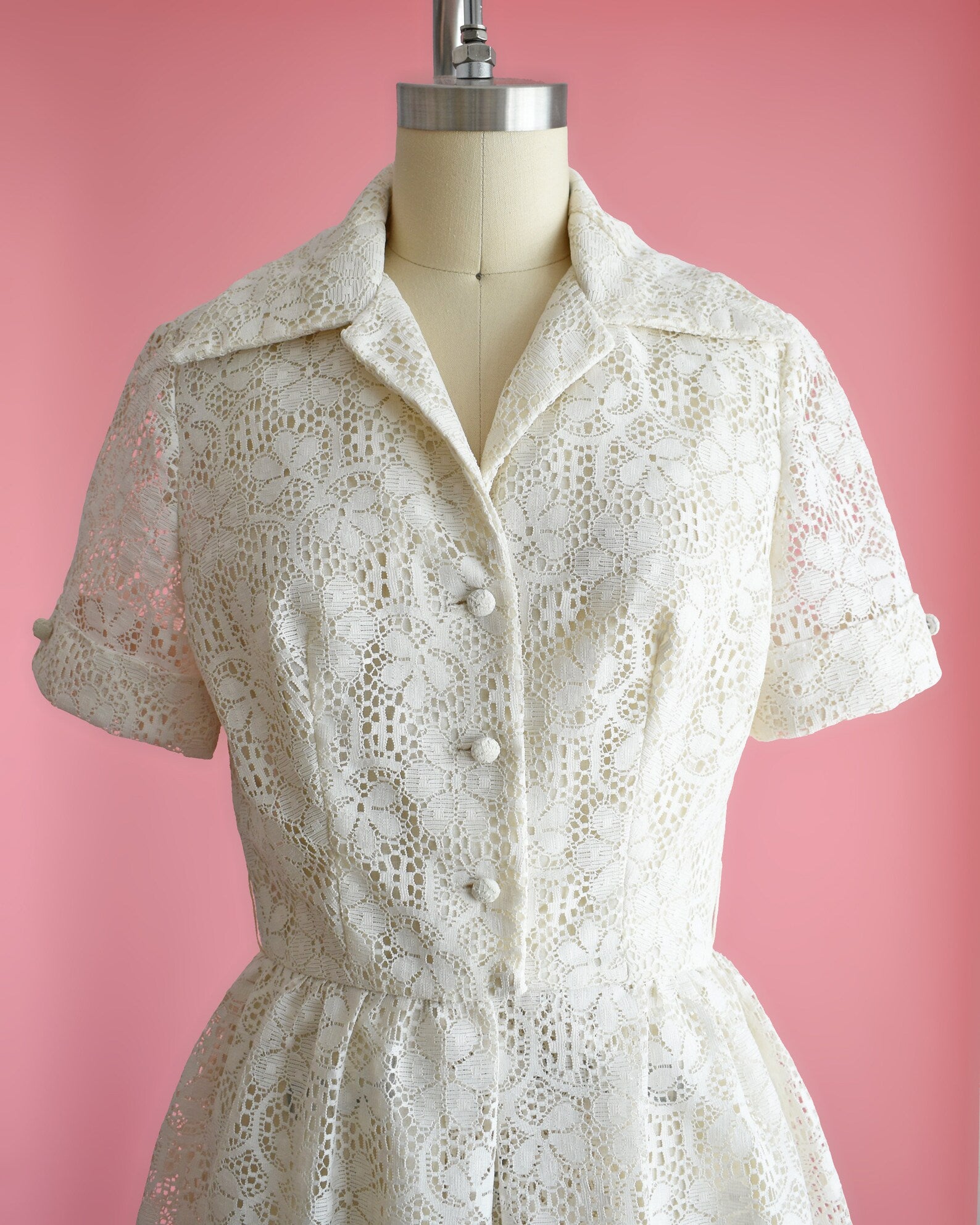 Close up of the bodice of a vintage cream floral lace dress with collared neckline, short sleeves, and button front on a dress form set on a pink background
