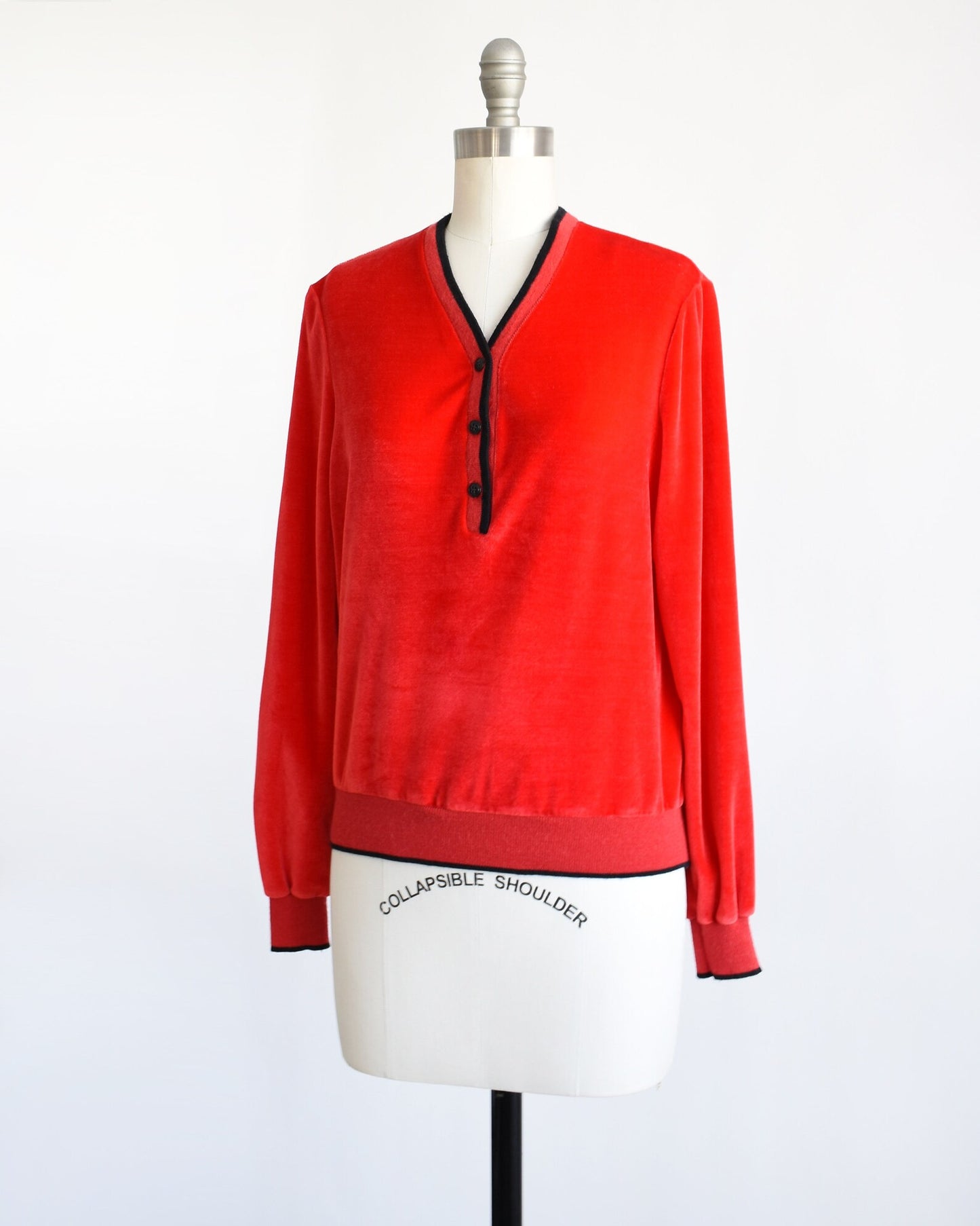 Side front view of a vintage 70s red velour top with black trim along the v-neckline, cuffs, and hem. There are three black buttons on the front and the top is modeled on a dress form.