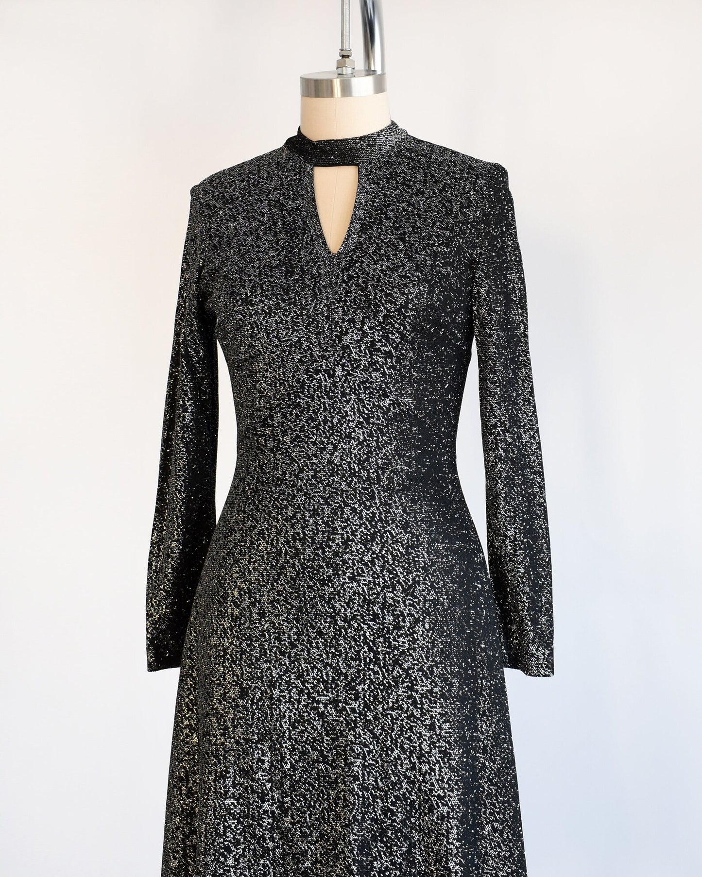 Side front view of a vintage 70s black and silver metallic dress that has an upside down triangle cutout under the front of the collar, long sleeves, and a knee length skirt. The dress is modeled on a dress form.