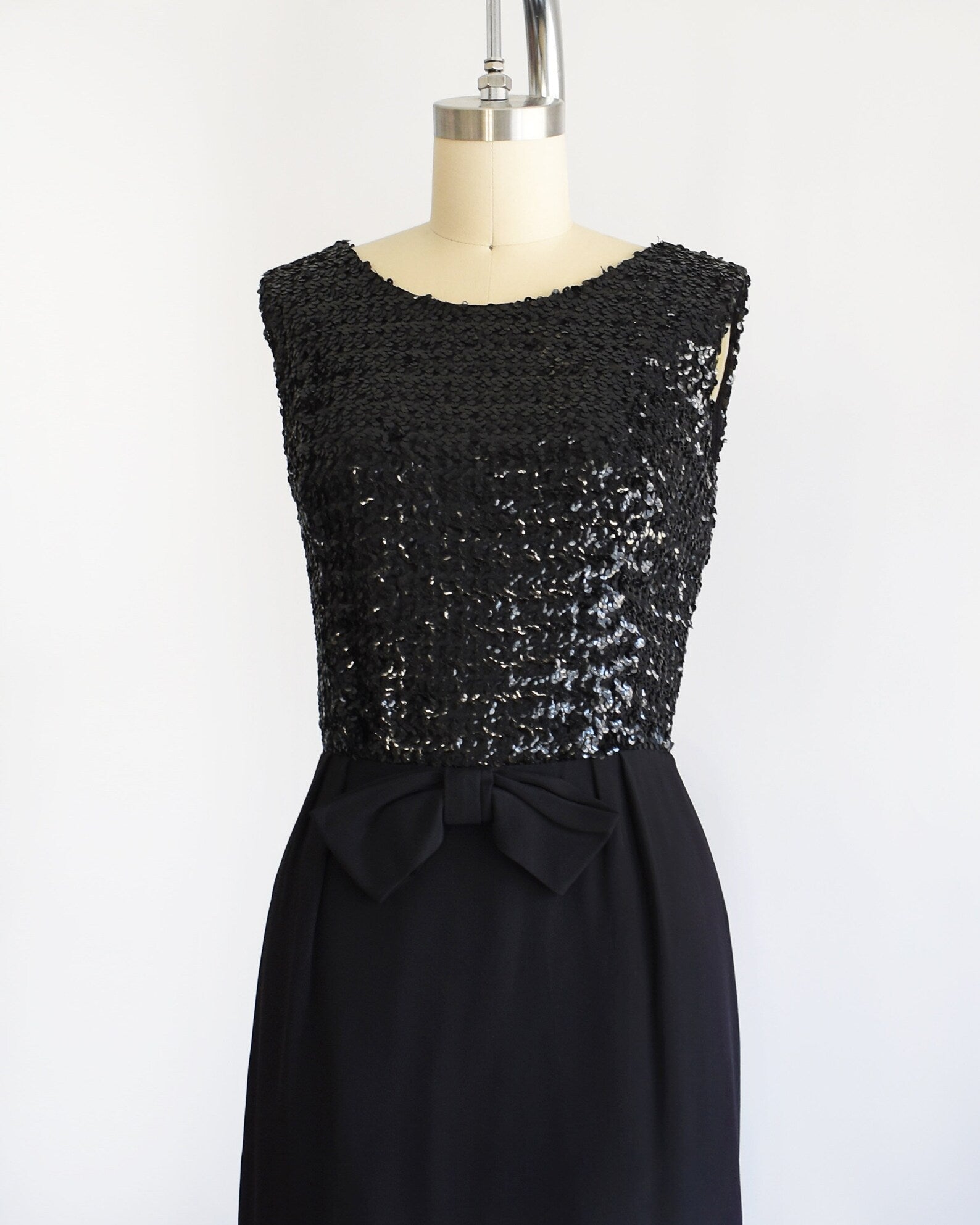 Side front view of a vintage late 50s early 60s black dress that has a black sequin bodice, a bow at the waist, and a pencil skirt. The dress is modeled on a dress form