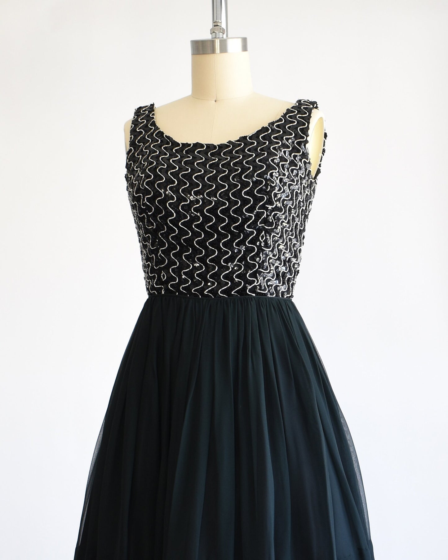 Side front view of a vintage 60s dress with a black sequin bodice with silver vertical squiggles. The skirt is a layered with black chiffon on top. The dress is modeled on a dress form.