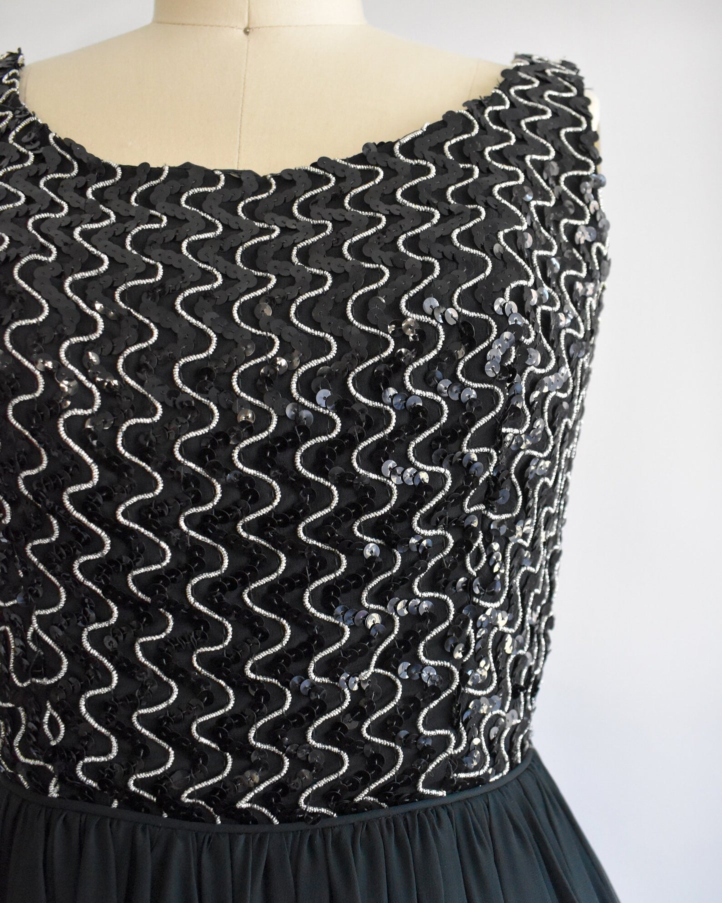 Close up of a  vintage 60s dress with a black sequin bodice with silver vertical squiggles. The skirt is a layered with black chiffon on top. The dress is modeled on a dress form.