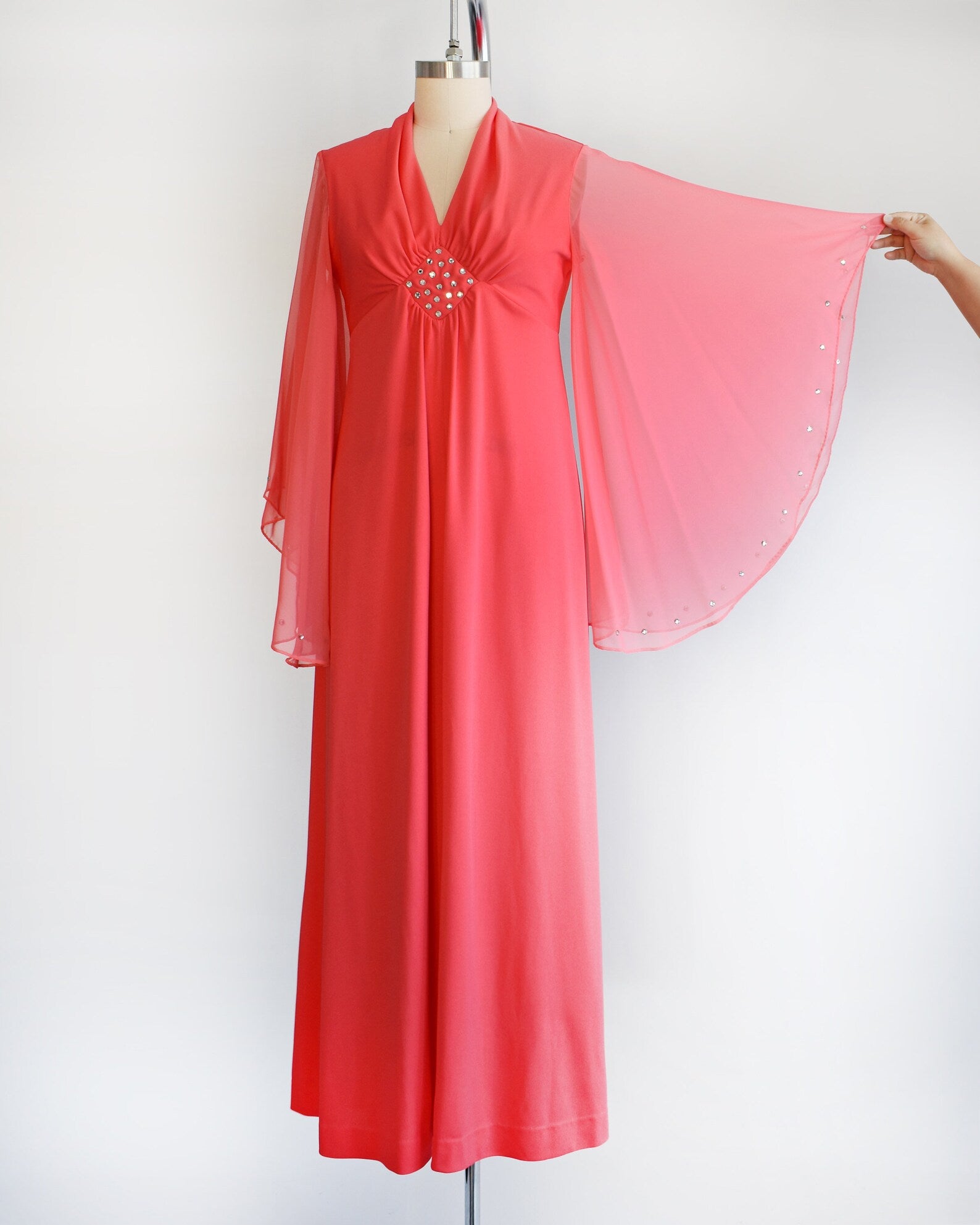 A vintage 70s coral pink maxi dress that has a v-neckline, rhinestones on the waist, and semi-sheer long angel sleeves with matching rhinestone trim. The dress is modeled on a dress form and is shown with the wide sweep on the sleeves