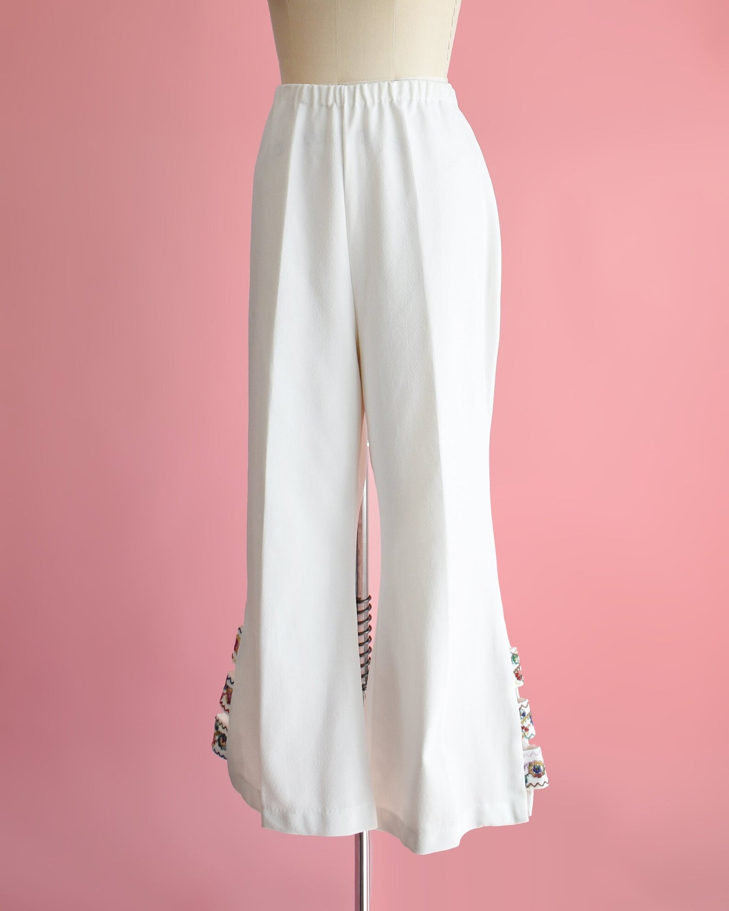 Side front view of a pair of vintage 70s white bell bottom pants that have embroidered cut out detail on the side of the cuffs. The pants are on a dress form.