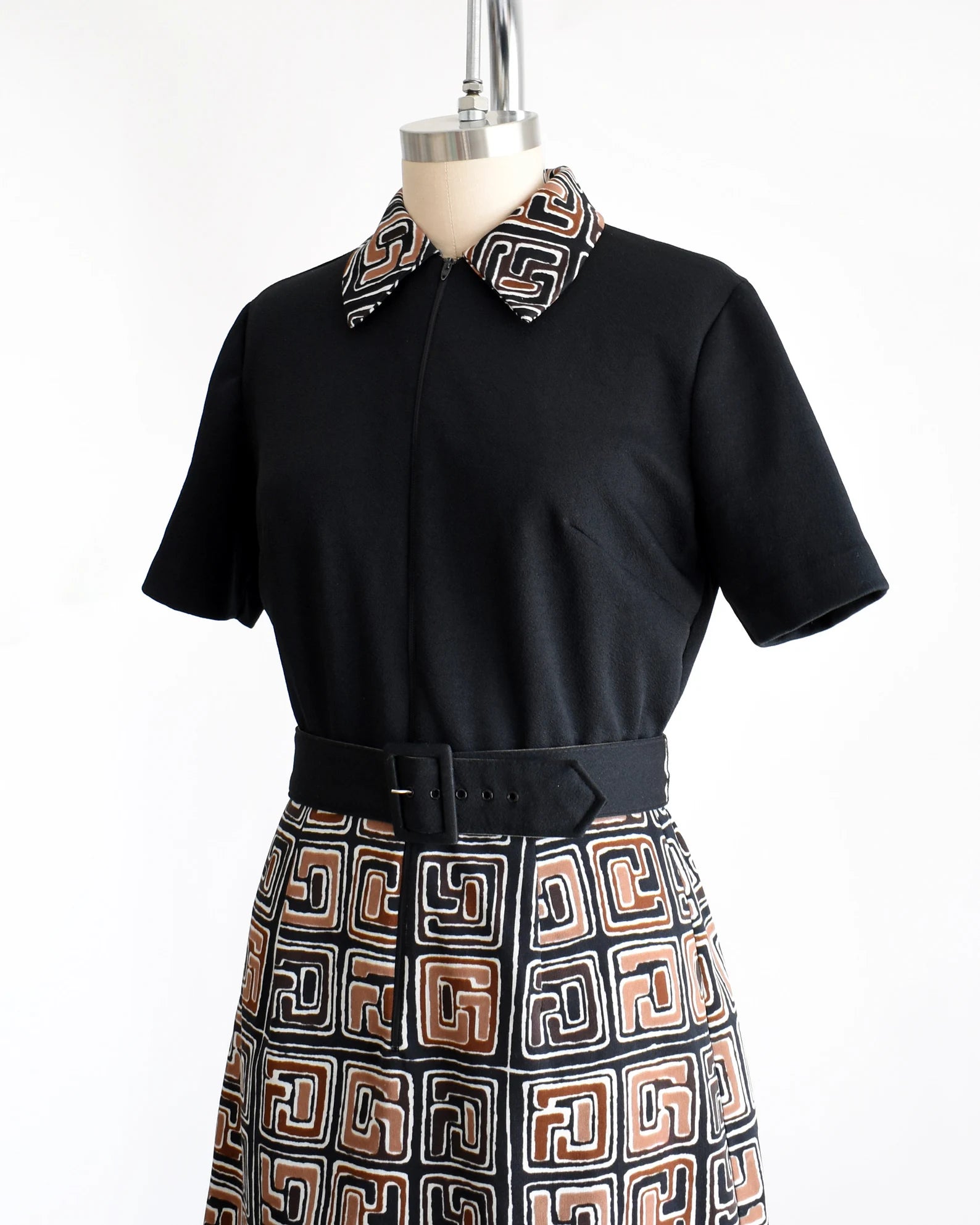 Side front view of a vintage 60s dress that has a matching geometric print brown, black and white collar and skirt. Zip up front. Matching black belt. Short sleeves. The dress is on a dress form.