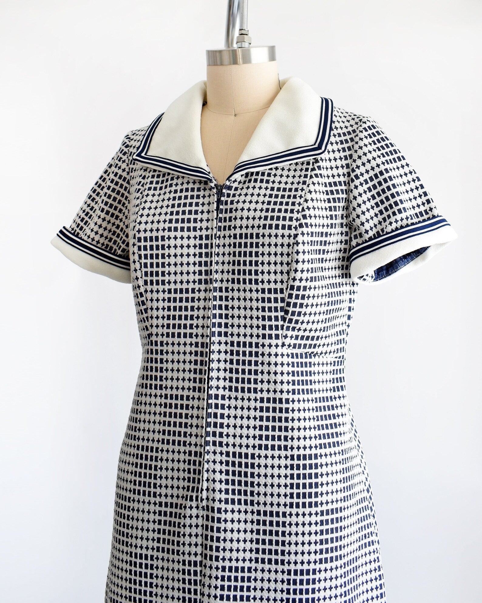 Side front view of a vintage 70s mod dress that has a blue and white geometric print, white wide collar with navy trim, zip up front, and matching white and navy trim on the short sleeves. The dress is on a dress form.