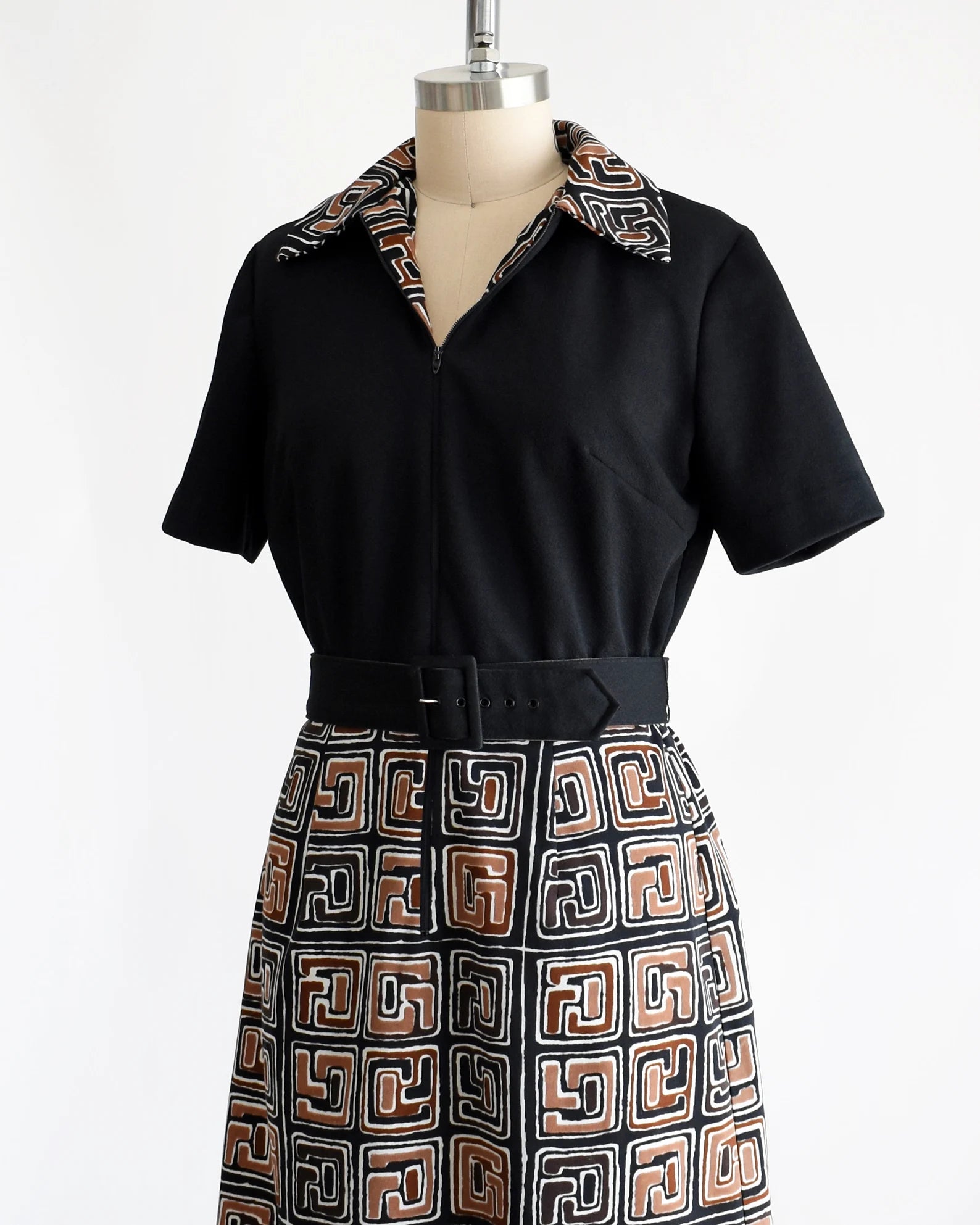 Side front view of a vintage 60s dress that has a matching geometric print brown, black and white collar and skirt. Zip up front that is zipped down a bit showing more of the collar. Matching black belt. Short sleeves. The dress is on a dress form.