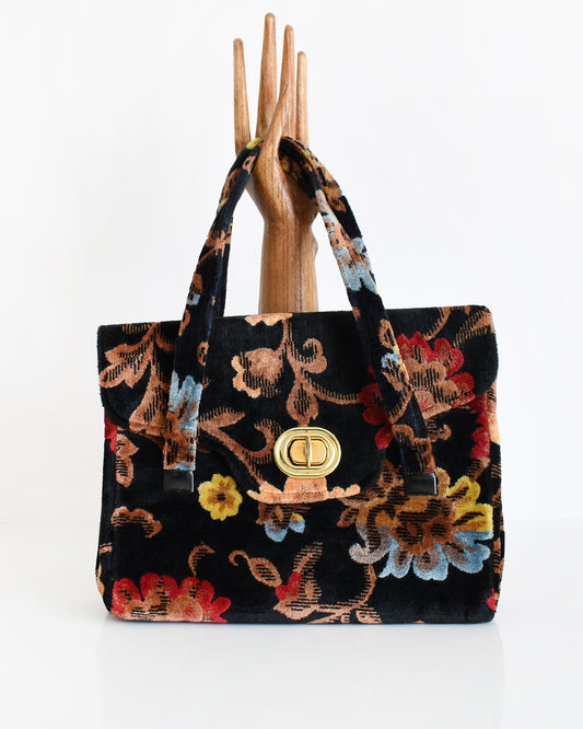 A vintage black floral velvet chenille handbag with gold tone hardware modeled with a wooden hand on a white table. The flowers are red, blue, yellow and have brown stems and leaves.