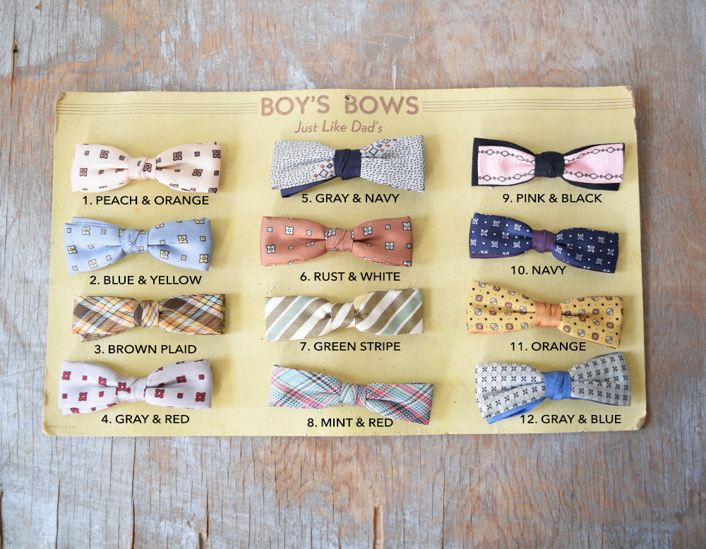 12 different types of clip on bow ties on all sorts of colors and prints on a yellow board. Each bow tie has a description of color underneath,