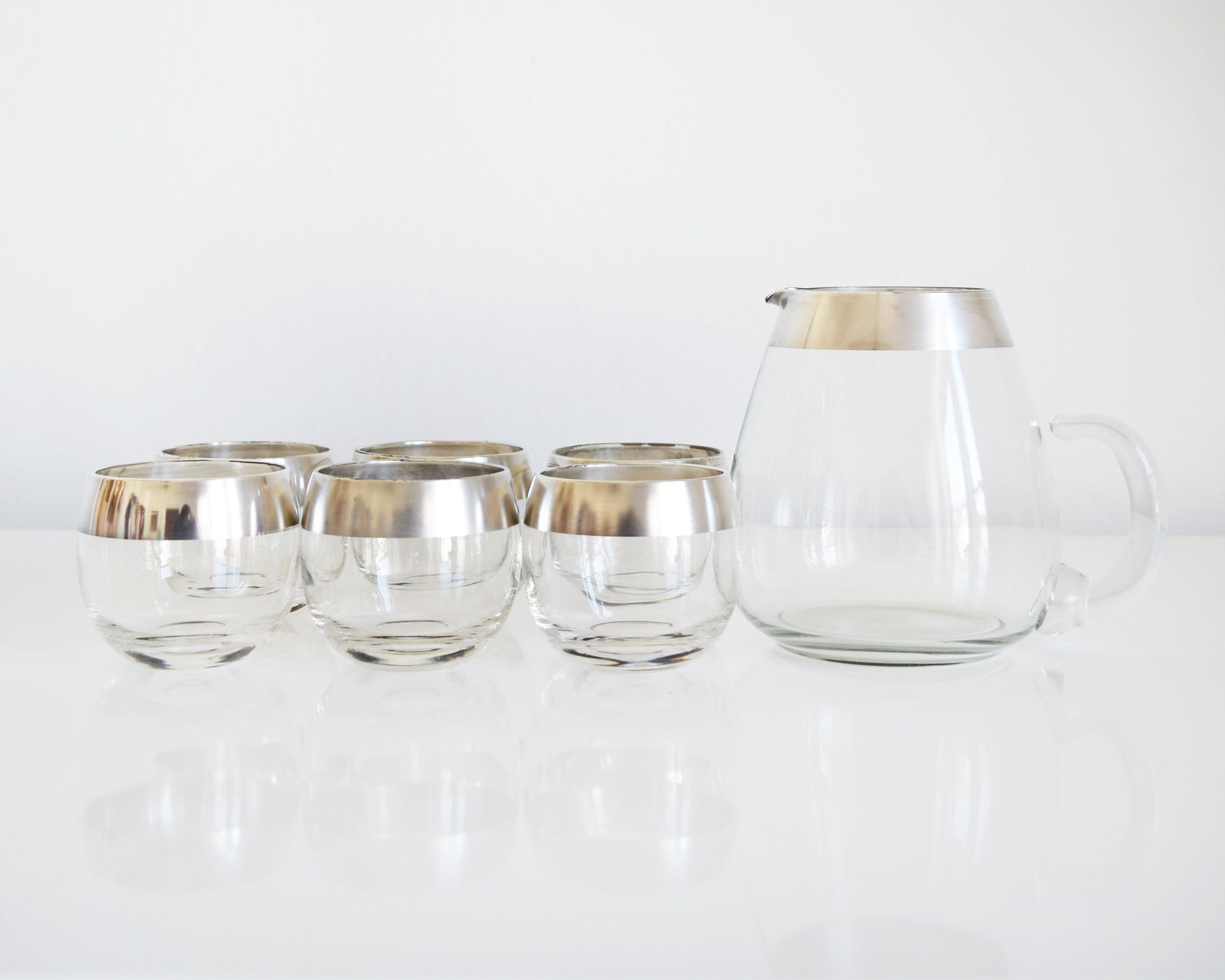 A vintage Dorothy Thorpe cocktail set that has a set of six silver rimmed rounded roly poly glasses with a matching silver rim pitcher with handle.
