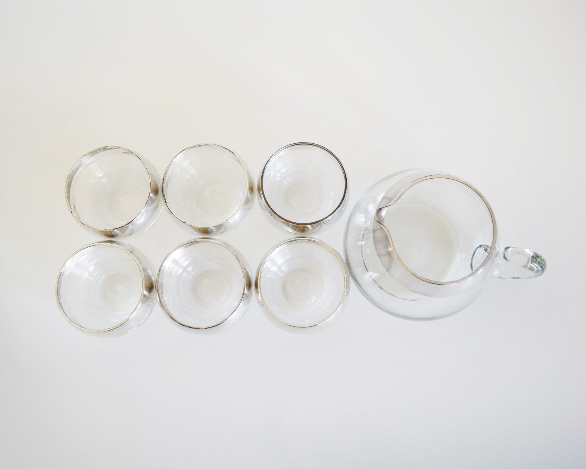 Overhead shot of a vintage Dorothy Thorpe cocktail set that has a set of six silver rimmed rounded roly poly glasses with a matching silver rim pitcher with handle.