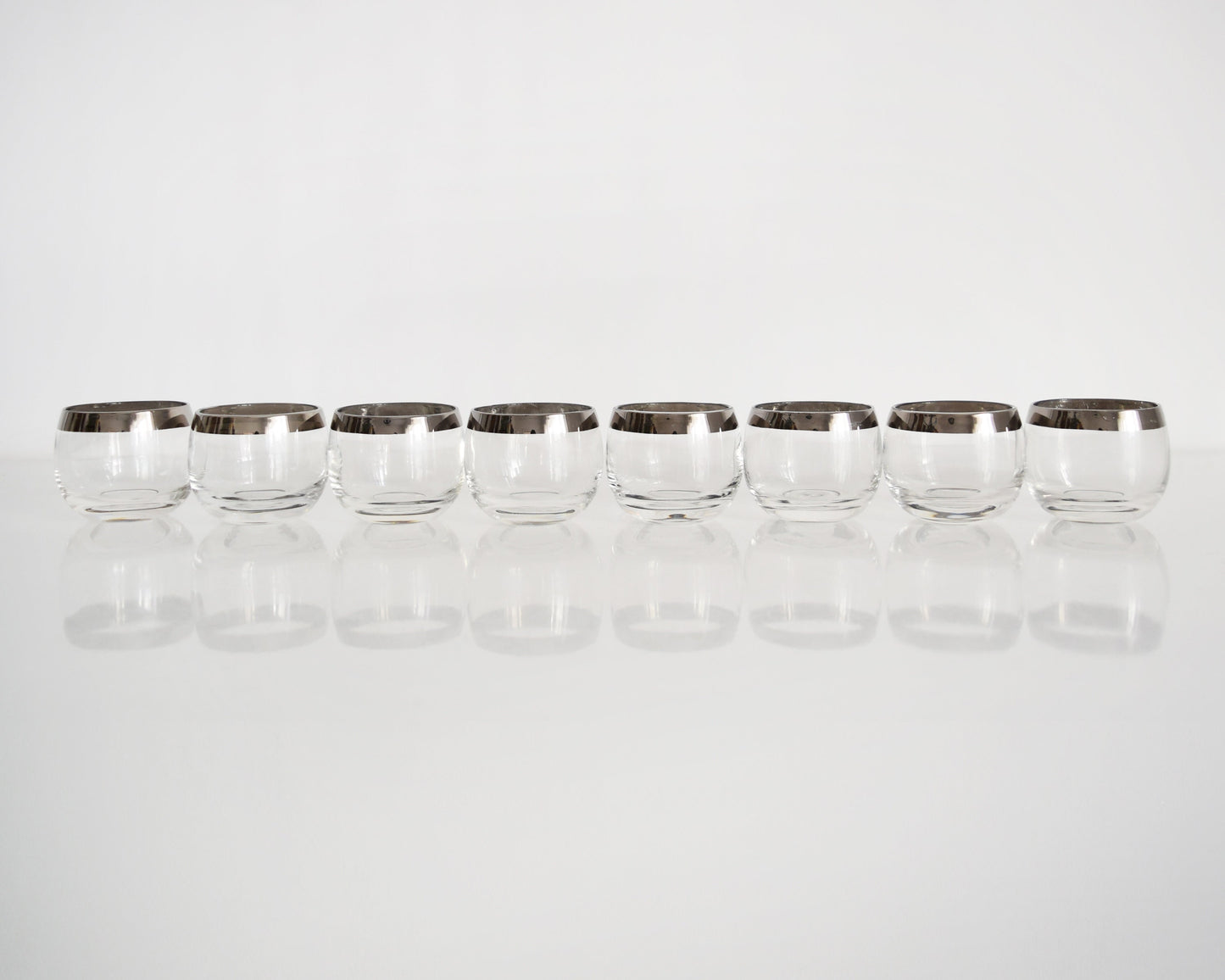 A row of eight silver rim roly poly glasses that are on a white table against a white background
