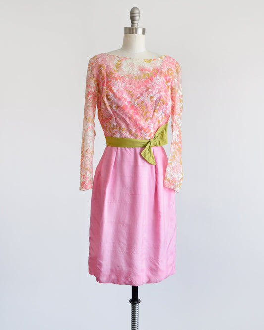 A vintage 60s dress that has a pink, green, and white sheer floral lace bodice. Long sleeves with small slits on the inside of the cuffs. Fitted waist that's adorned with a green bow sash belt which hooks on the back. 
