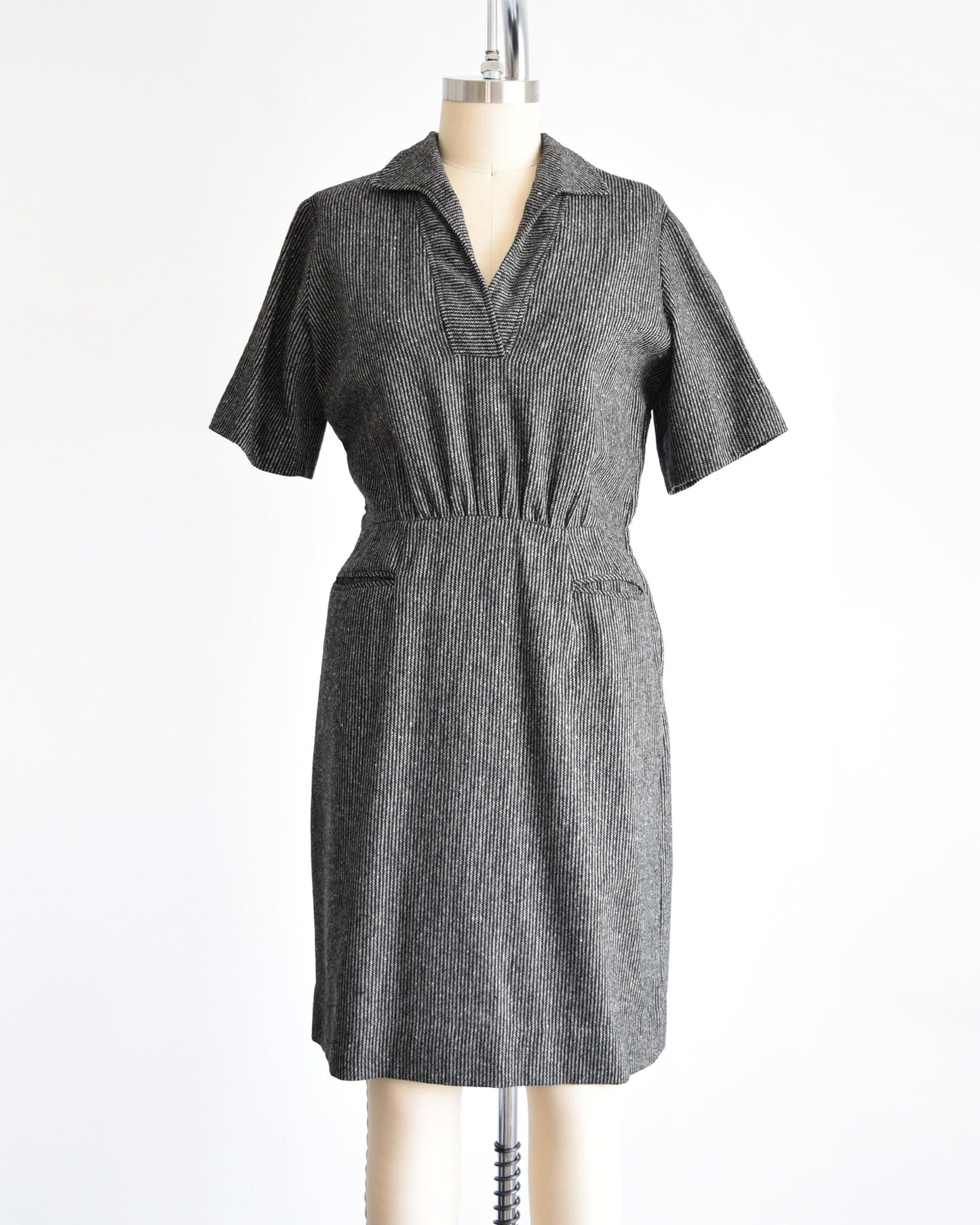 A 1950s vintage wool dress that is black and white striped with a v-neck collared neckline, short sleeves, fitted waist, and two front pockets below the waist. Dress is on a dress form