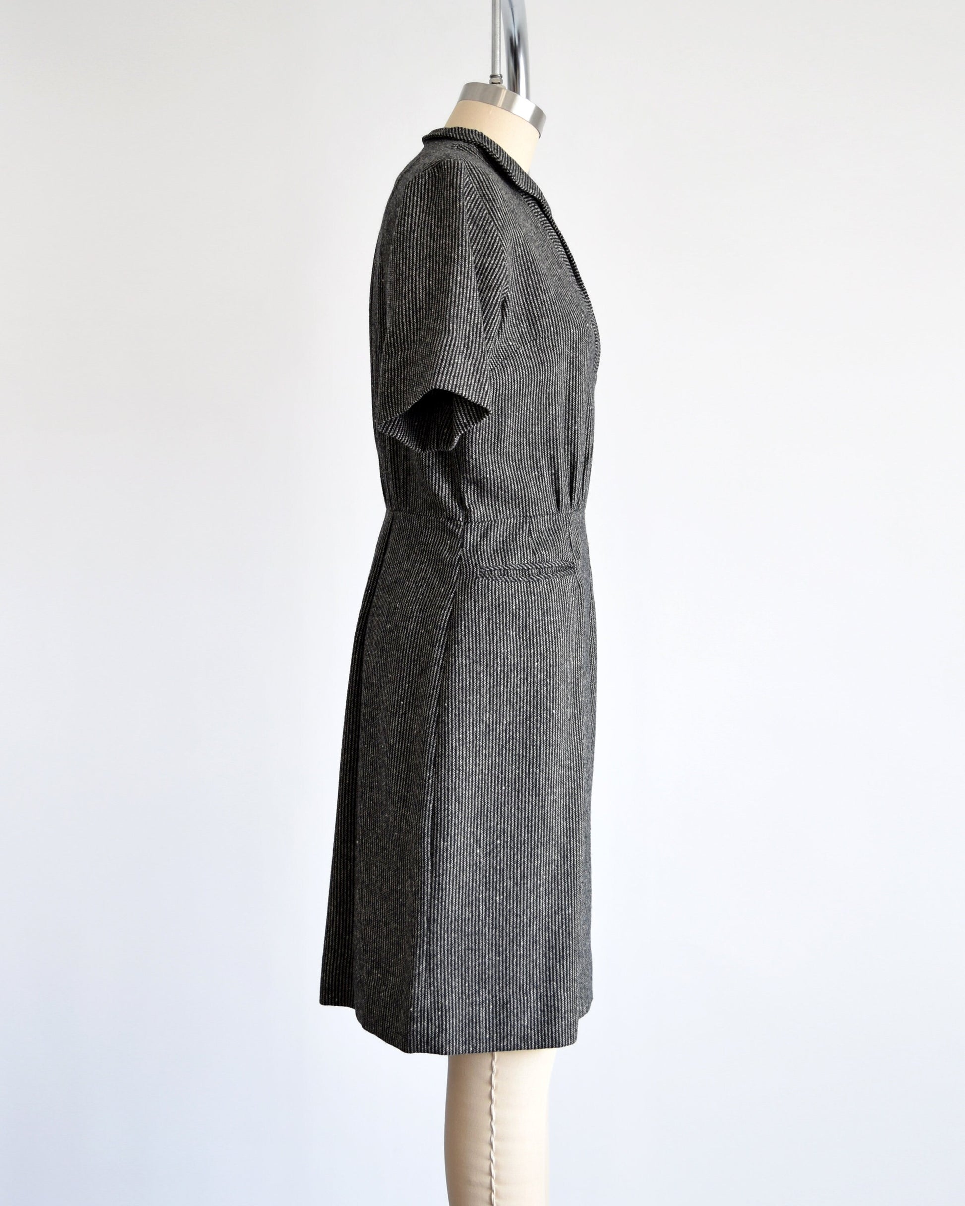 Side view of a 1950s vintage wool dress that is black and white striped with a v-neck collared neckline, short sleeves, fitted waist, and two front pockets below the waist. Dress is on a dress form