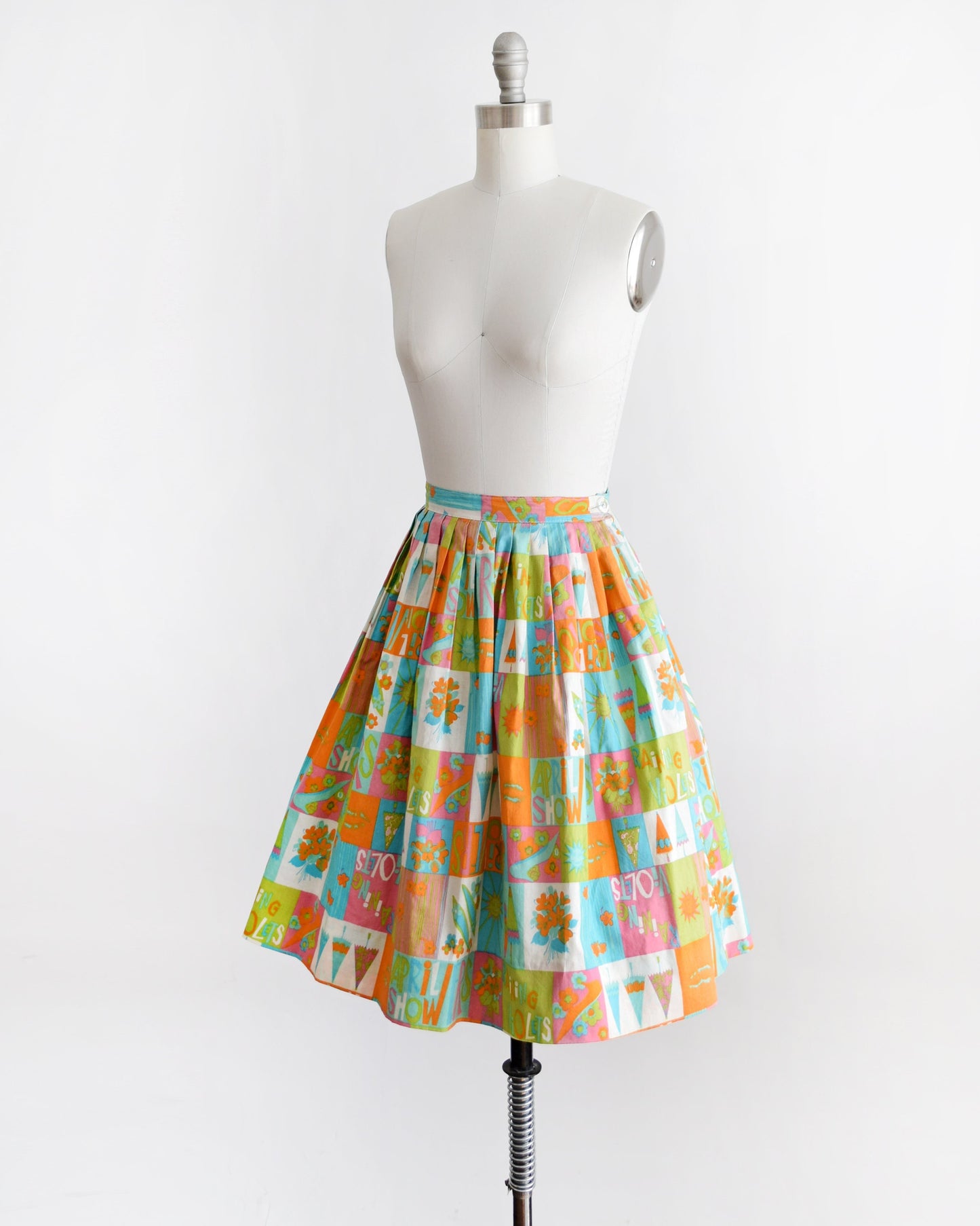 Side front view of a vintage 50s skirt has an April showers theme with colorful blocks of flowers, umbrellas, shoes, the sun, and the words April Showers and Raining Violets in a classic mid century print. Skirt is modeled on a dress form