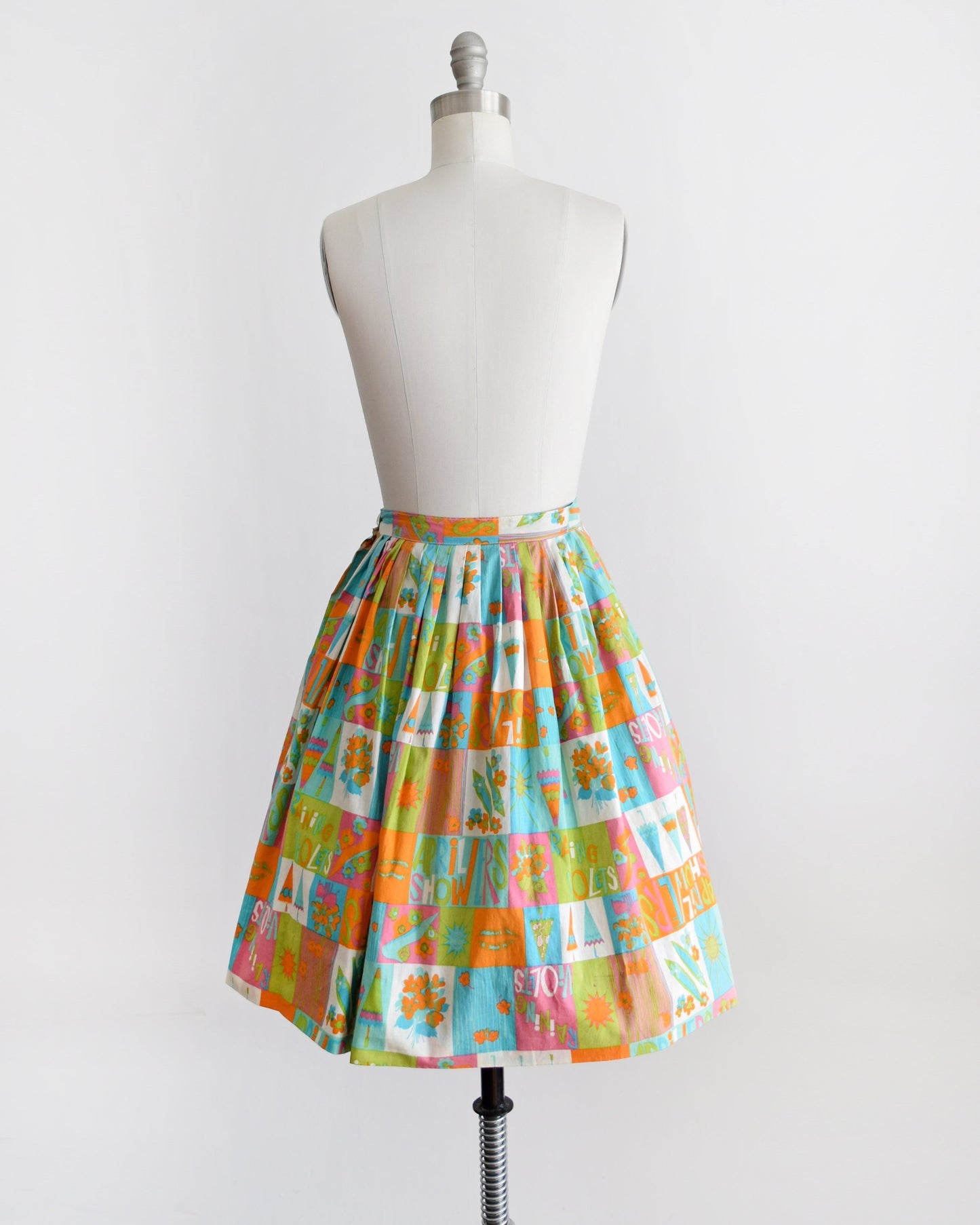 Back view of a vintage 50s skirt has an April showers theme with colorful blocks of flowers, umbrellas, shoes, the sun, and the words April Showers and Raining Violets in a classic mid century print. Skirt is modeled on a dress form