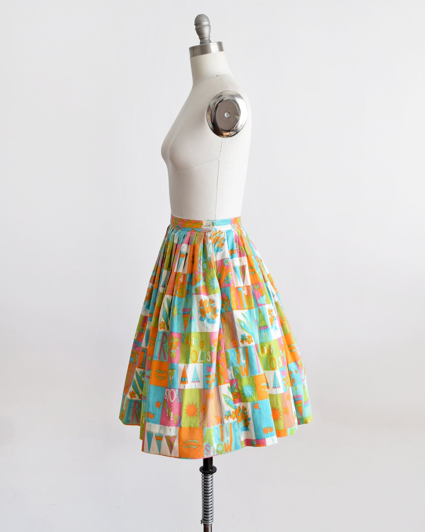 Side view of a vintage 50s skirt has an April showers theme with colorful blocks of flowers, umbrellas, shoes, the sun, and the words April Showers and Raining Violets in a classic mid century print. Skirt is modeled on a dress form
