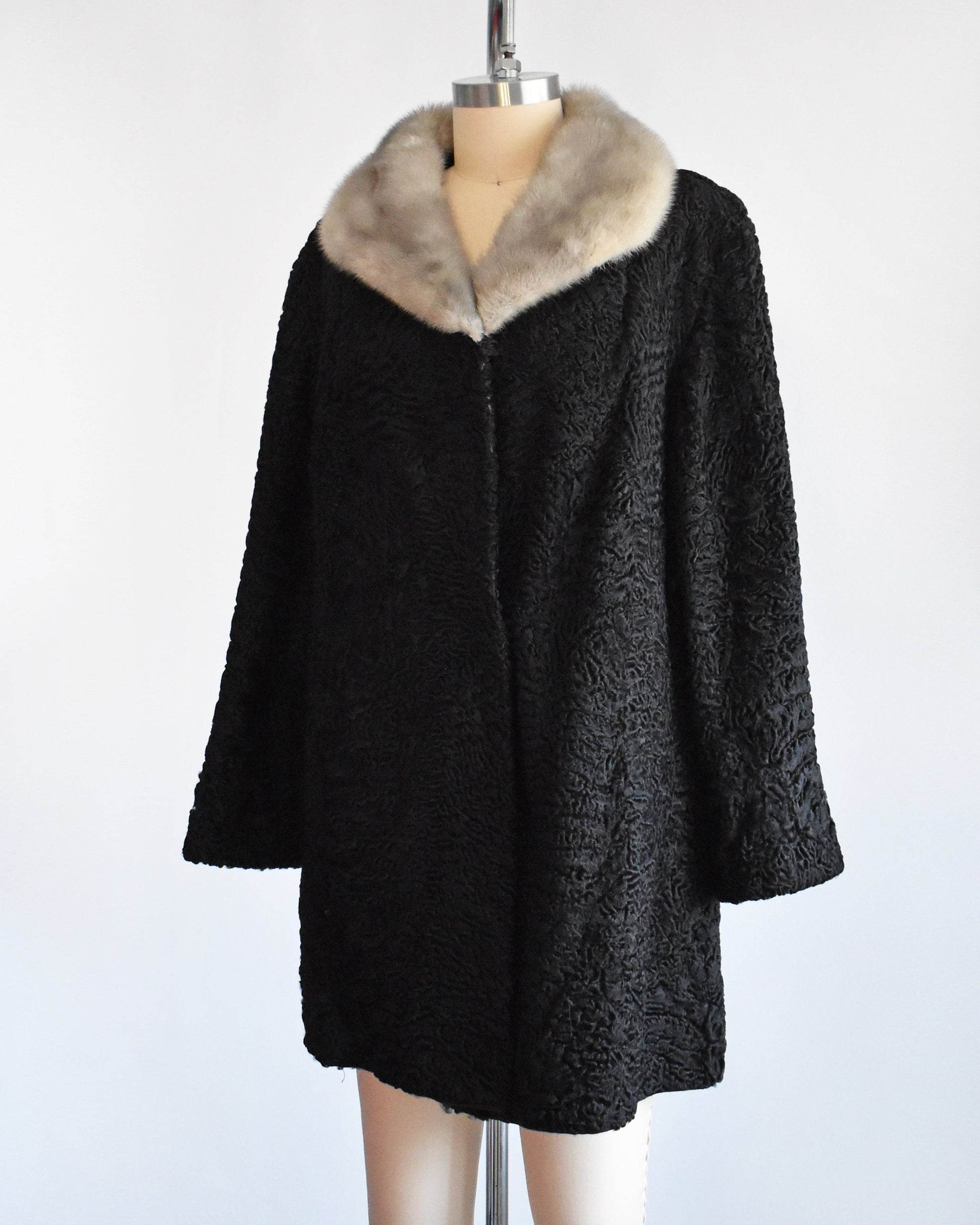 Side front view of a vintage late 50s early 60s black curly Persian lamb coat that has a silver gray mink fur collar. Two hidden hook and eye closures down the front. Coat is on a dress form.
