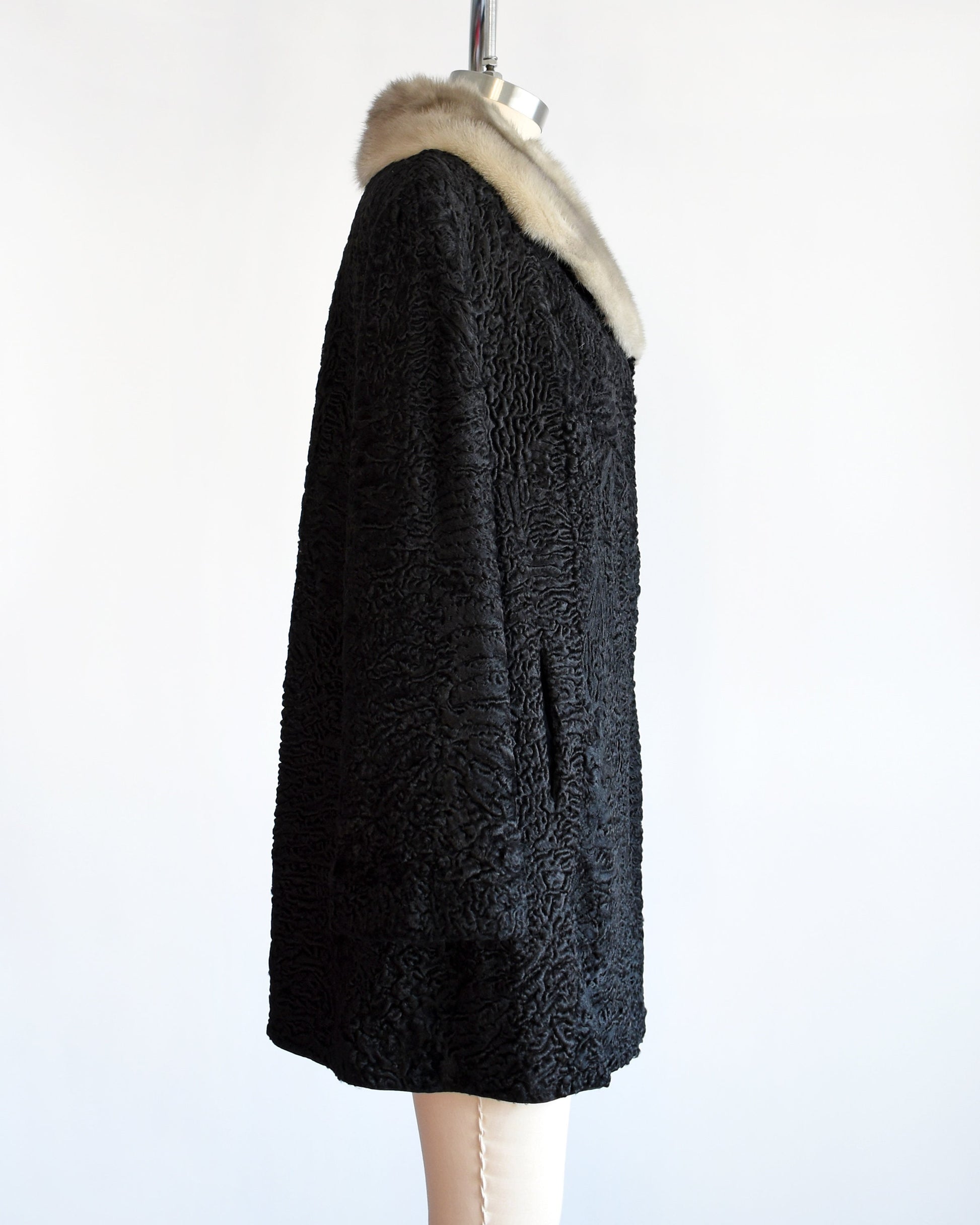 Side view of a vintage late 50s early 60s black curly Persian lamb coat that has a silver gray mink fur collar. Two hidden hook and eye closures down the front. Coat is on a dress form.
