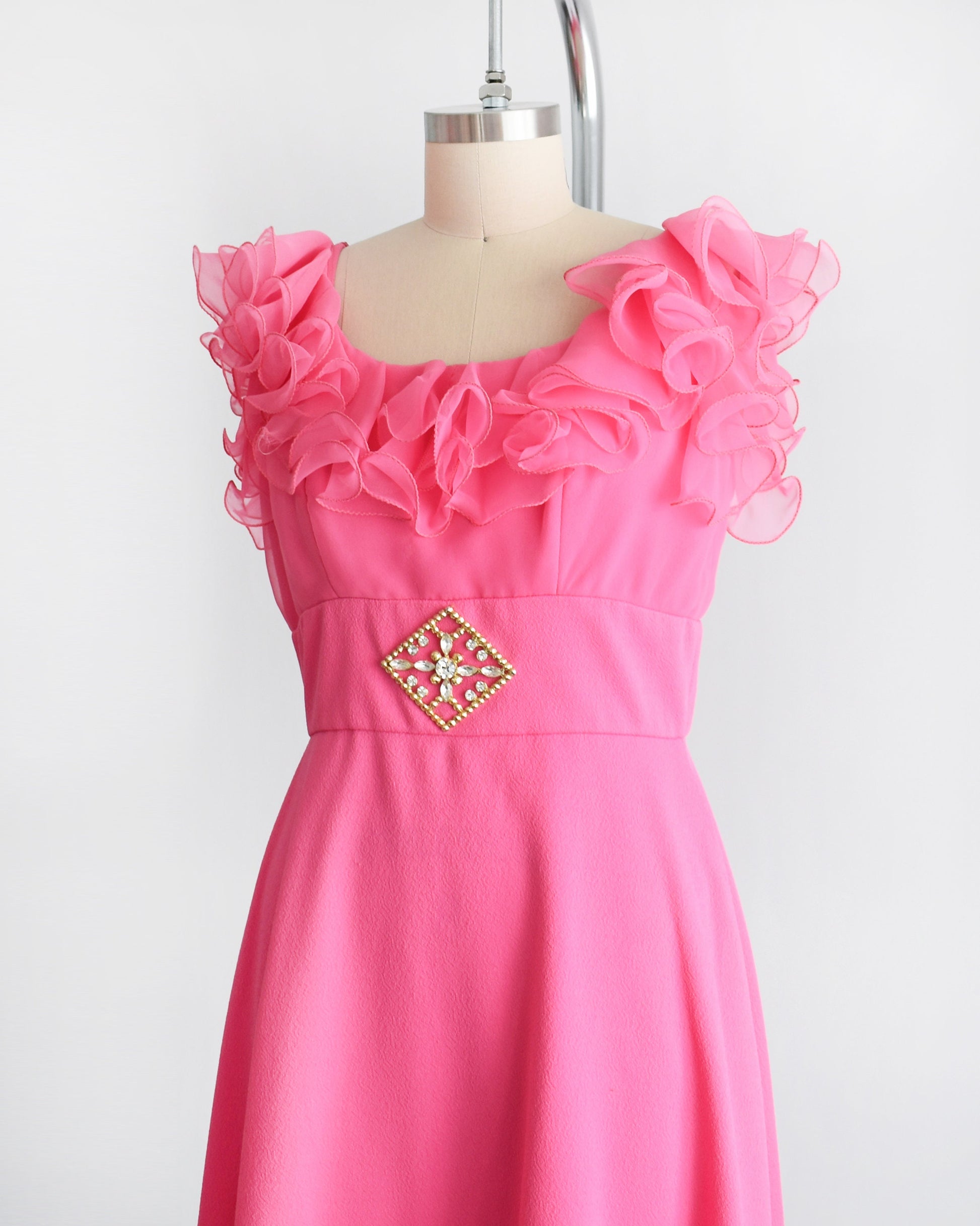 Side front view of a vintage 1970s hot pink maxi dress that has a ruffled neckline and a gold tone rhinestone brooch at the waist.