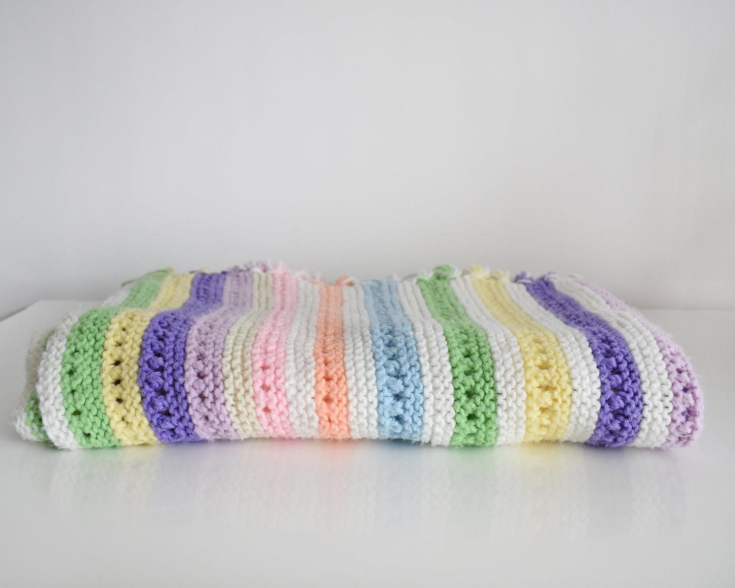 A white with dark and light purple, pink, blue, green, yellow, cream, and orange striped fringed blanket folded up on a white table.