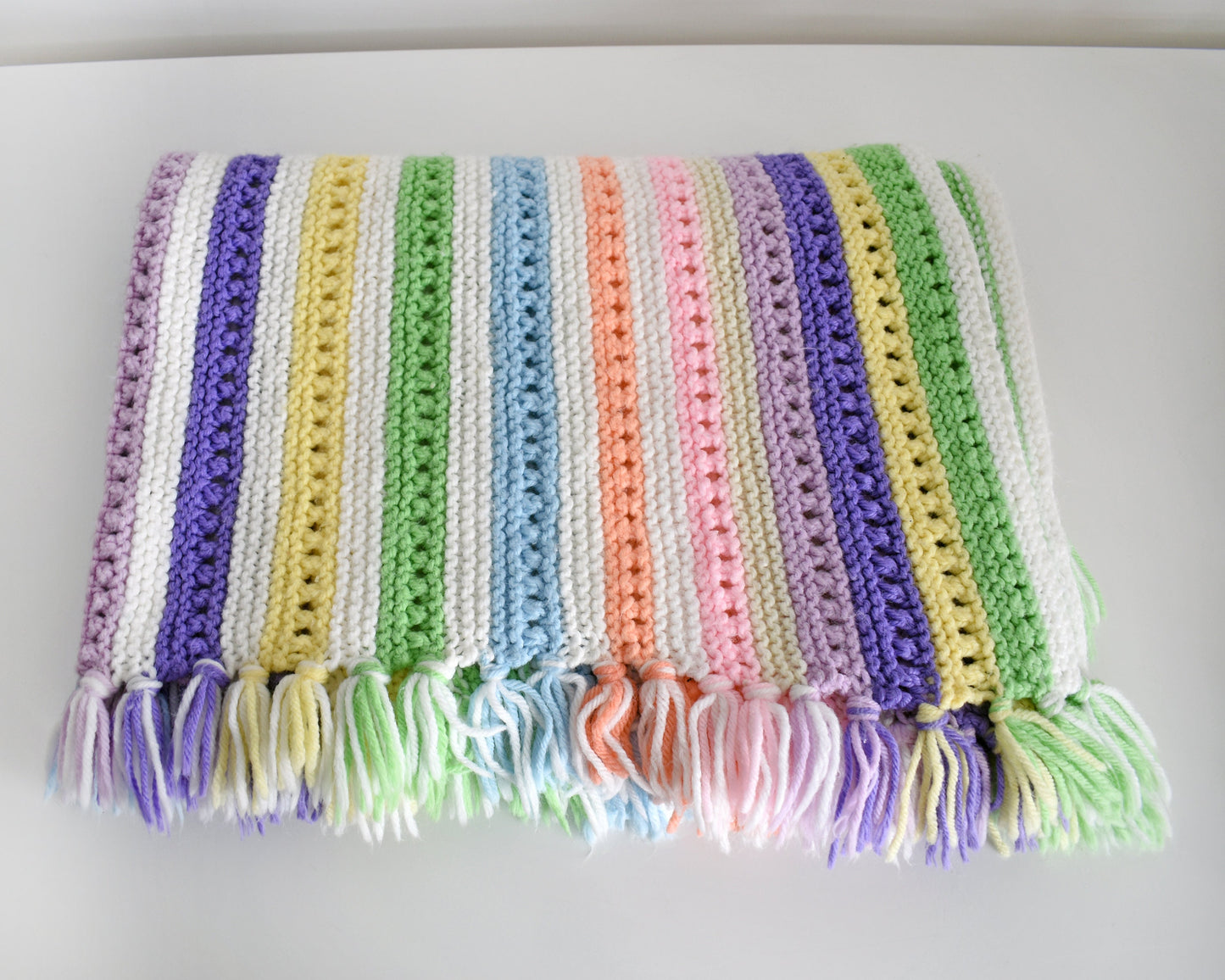 A white with dark and light purple, pink, blue, green, yellow, cream, and orange striped fringed blanket folded up on a white table.