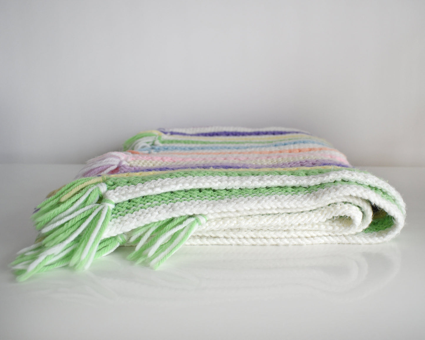 A white with dark and light purple, pink, blue, green, yellow, cream, and orange striped fringed blanket folded up on a white table.
