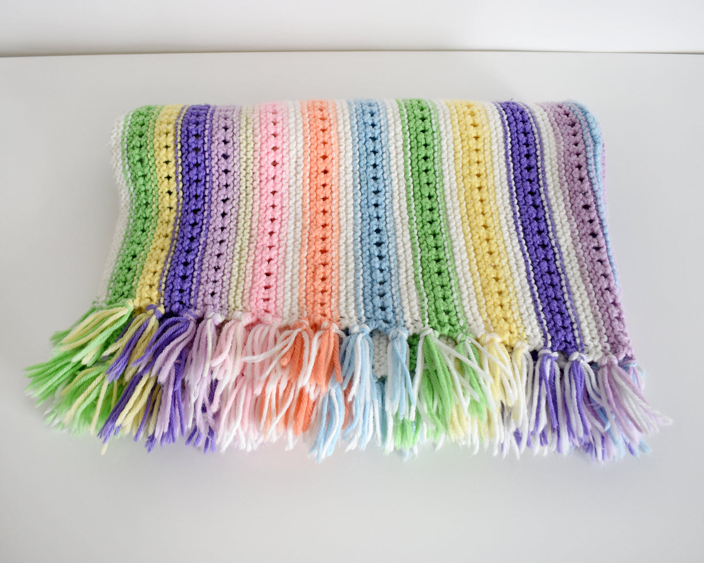 A white with dark and light purple, pink, blue, green, yellow, cream, and orange striped fringed blanket folded up on a white table.