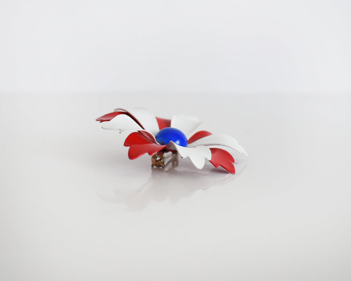 Side view of a vintage 60s mod floral brooch that has red and white petals with a blue rounded center. The pin is on a table