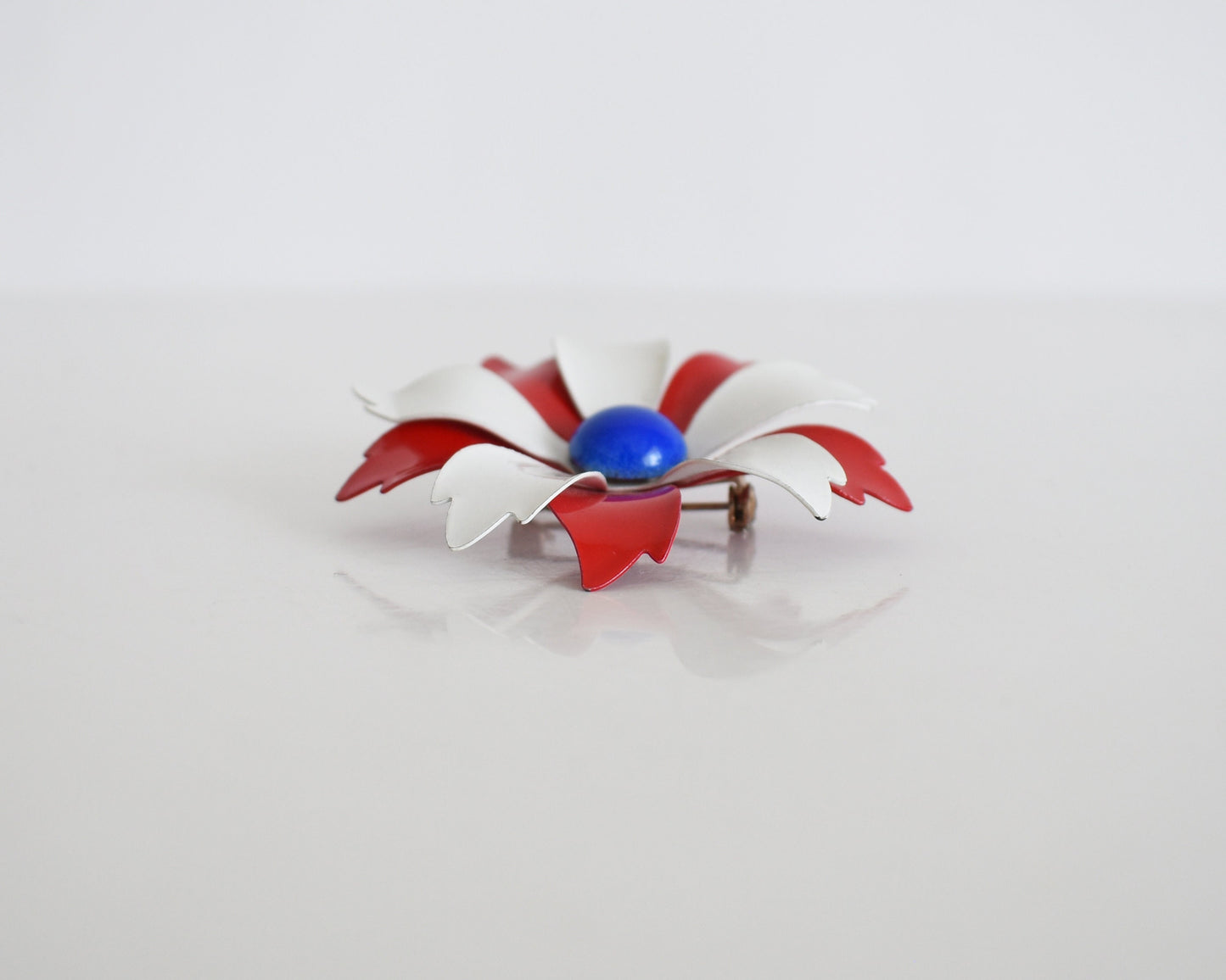 A vintage 60s mod floral brooch that has red and white petals with a blue rounded center. White and red backing with pin closure. The pin is on a table