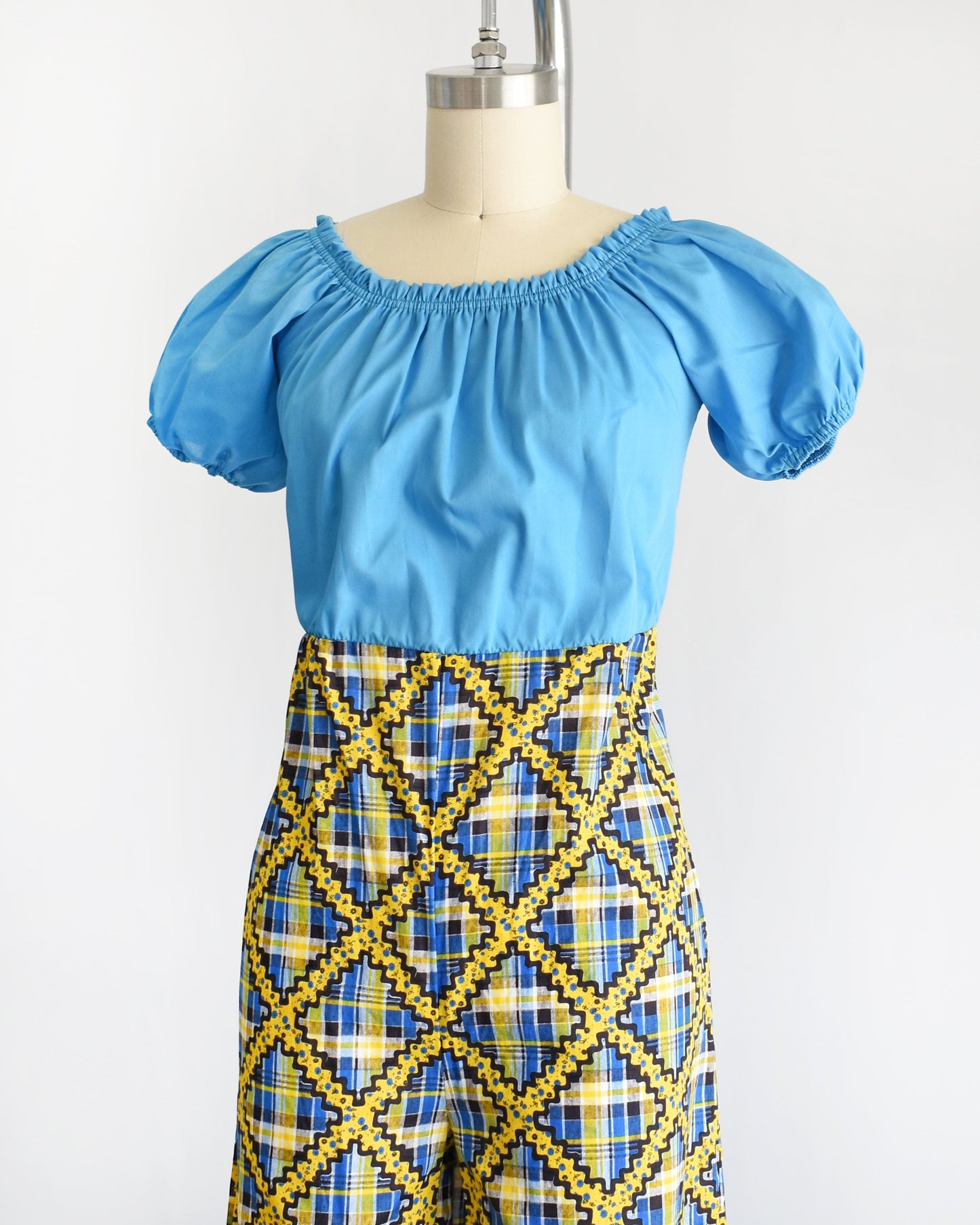 Side front view of a vintage 70s jumpsuit that has a blue bodice with smocked elastic neckline and puff sleeves. The pants are a blue, black, and yellow plaid, with a ric-rac dotted yellow and black print on top of the plaid.