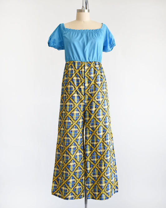 A vintage 70s jumpsuit that has a blue bodice with smocked elastic neckline and puff sleeves. The pants are a blue, black, and yellow plaid, with a ric-rac dotted yellow and black print on top of the plaid.