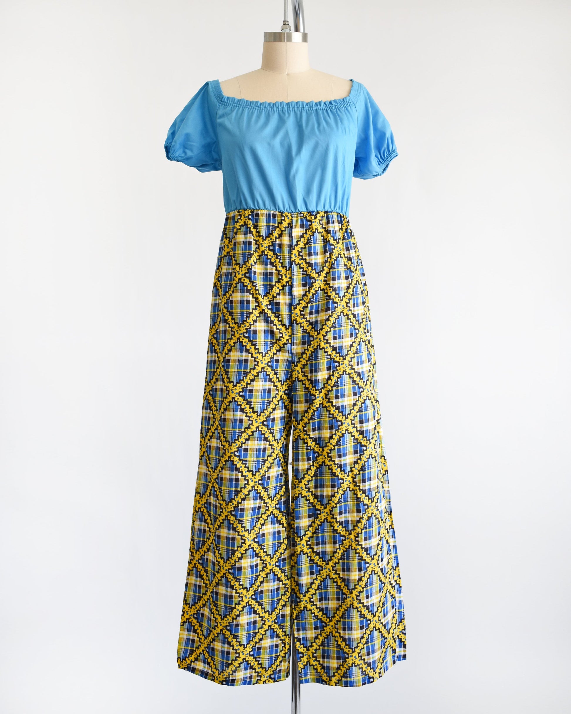 A vintage 70s jumpsuit that has a blue bodice with smocked elastic neckline and puff sleeves. The pants are a blue, black, and yellow plaid, with a ric-rac dotted yellow and black print on top of the plaid.