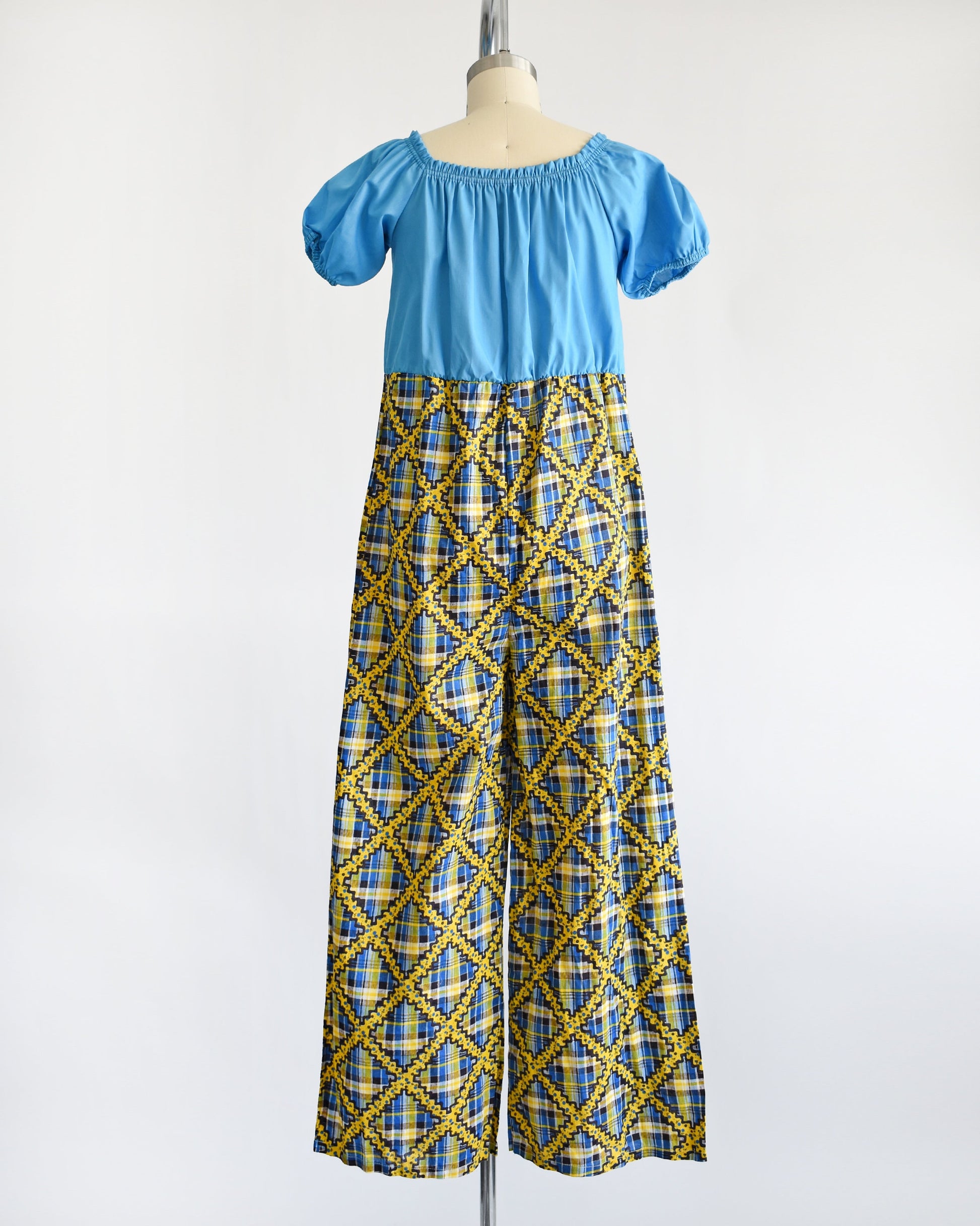 Back view of a vintage 70s jumpsuit that has a blue bodice with smocked elastic neckline and puff sleeves. The pants are a blue, black, and yellow plaid, with a ric-rac dotted yellow and black print on top of the plaid.
