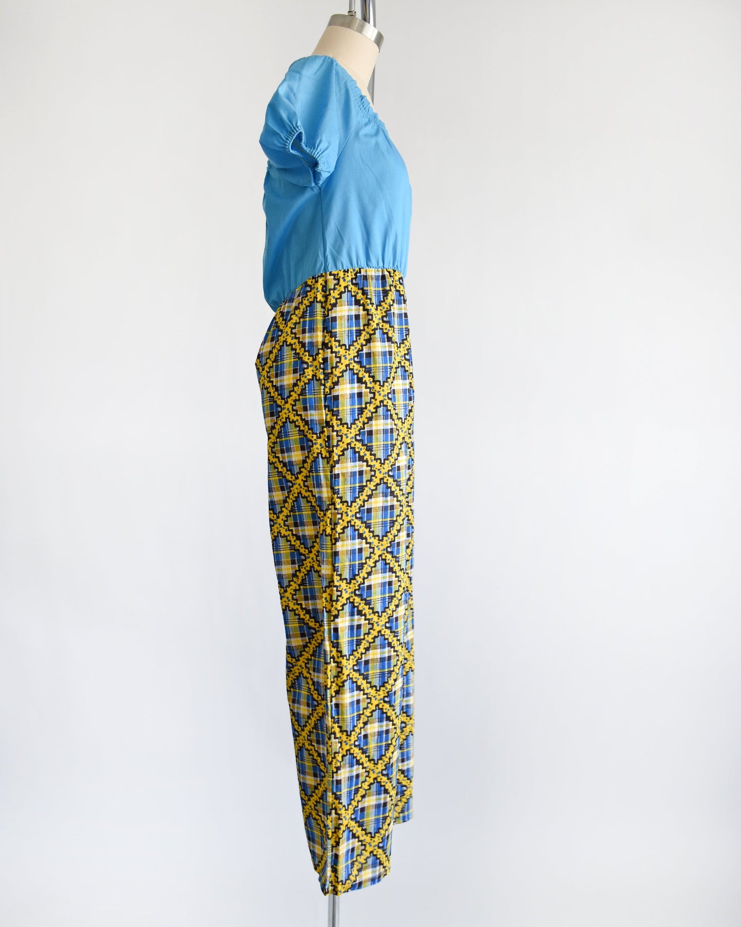 Side view of a vintage 70s jumpsuit that has a blue bodice with smocked elastic neckline and puff sleeves. The pants are a blue, black, and yellow plaid, with a ric-rac dotted yellow and black print on top of the plaid.