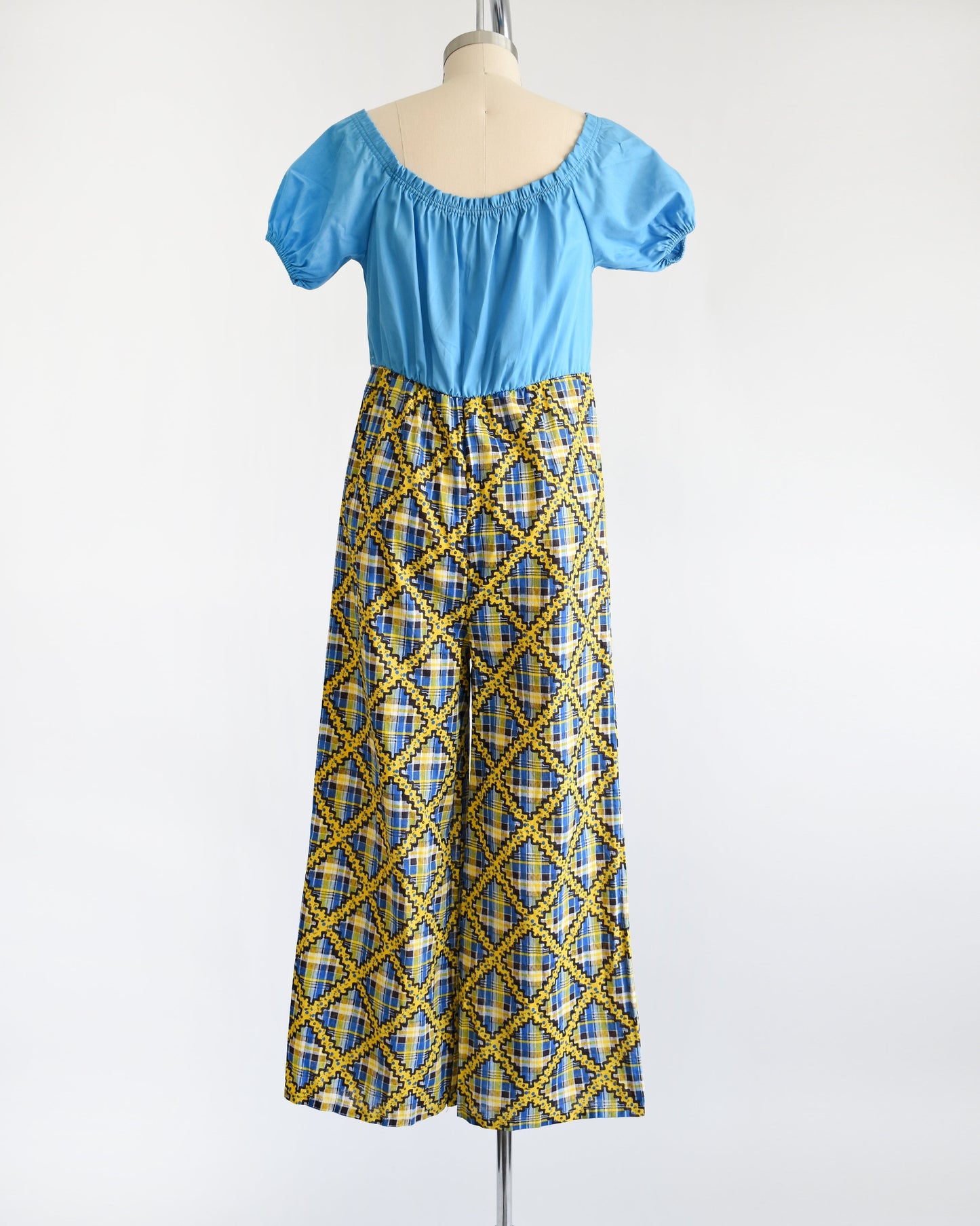 Back view of a vintage 70s jumpsuit that has a blue bodice with smocked elastic neckline and puff sleeves. The pants are a blue, black, and yellow plaid, with a ric-rac dotted yellow and black print on top of the plaid.