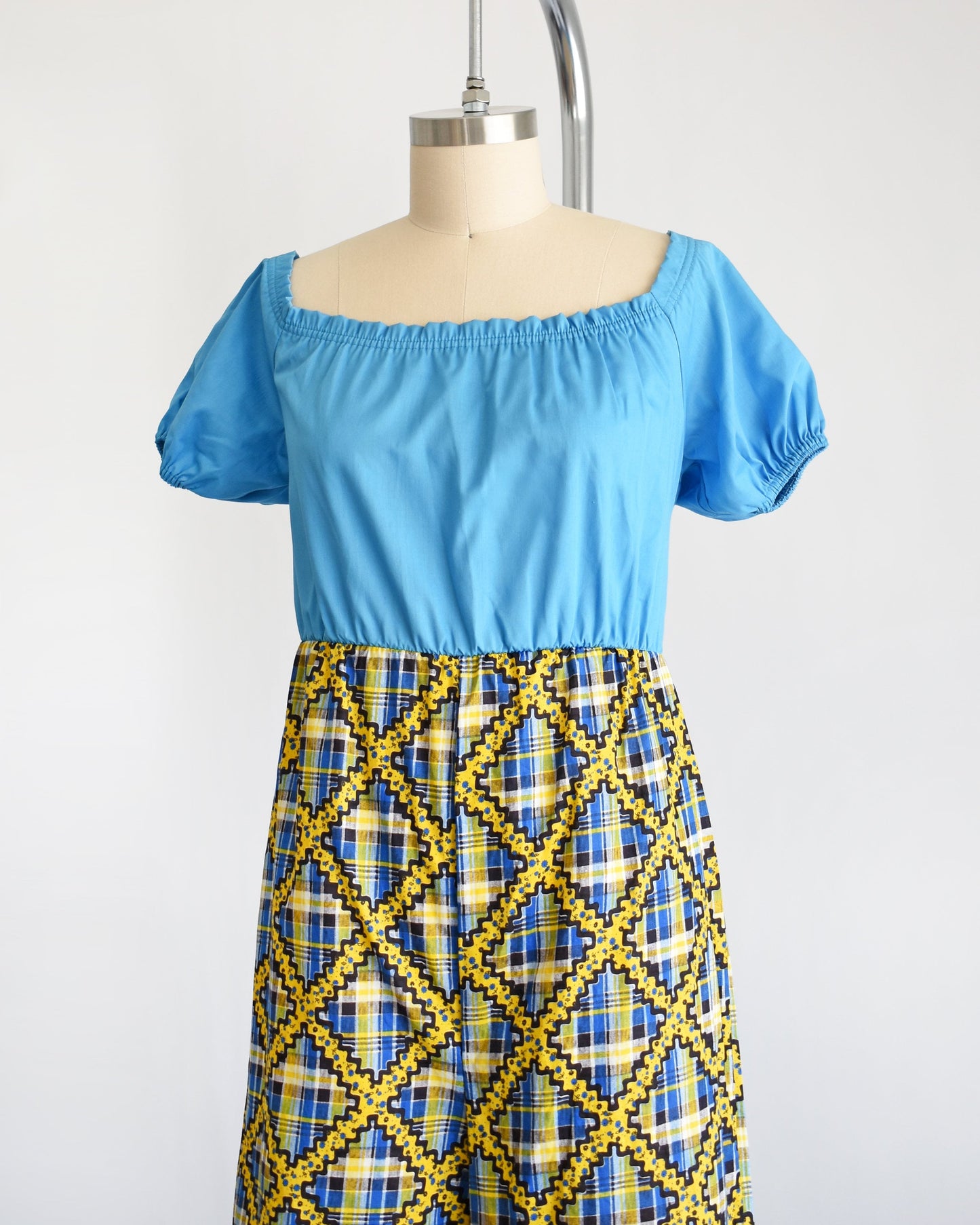 Side front view of a vintage 70s jumpsuit that has a blue bodice with smocked elastic neckline and puff sleeves. The pants are a blue, black, and yellow plaid, with a ric-rac dotted yellow and black print on top of the plaid.