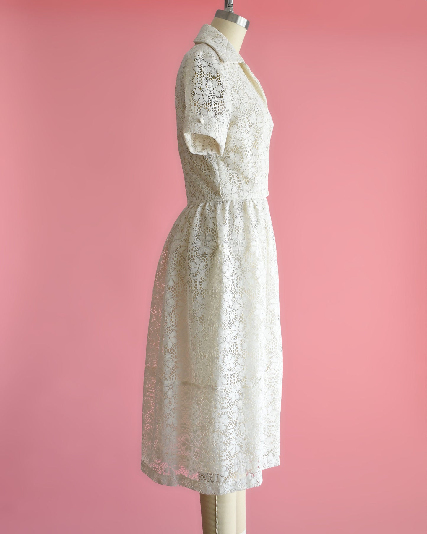 Side view of a vintage cream floral lace dress with collared neckline, short sleeves, and button front on a dress form set on a pink background