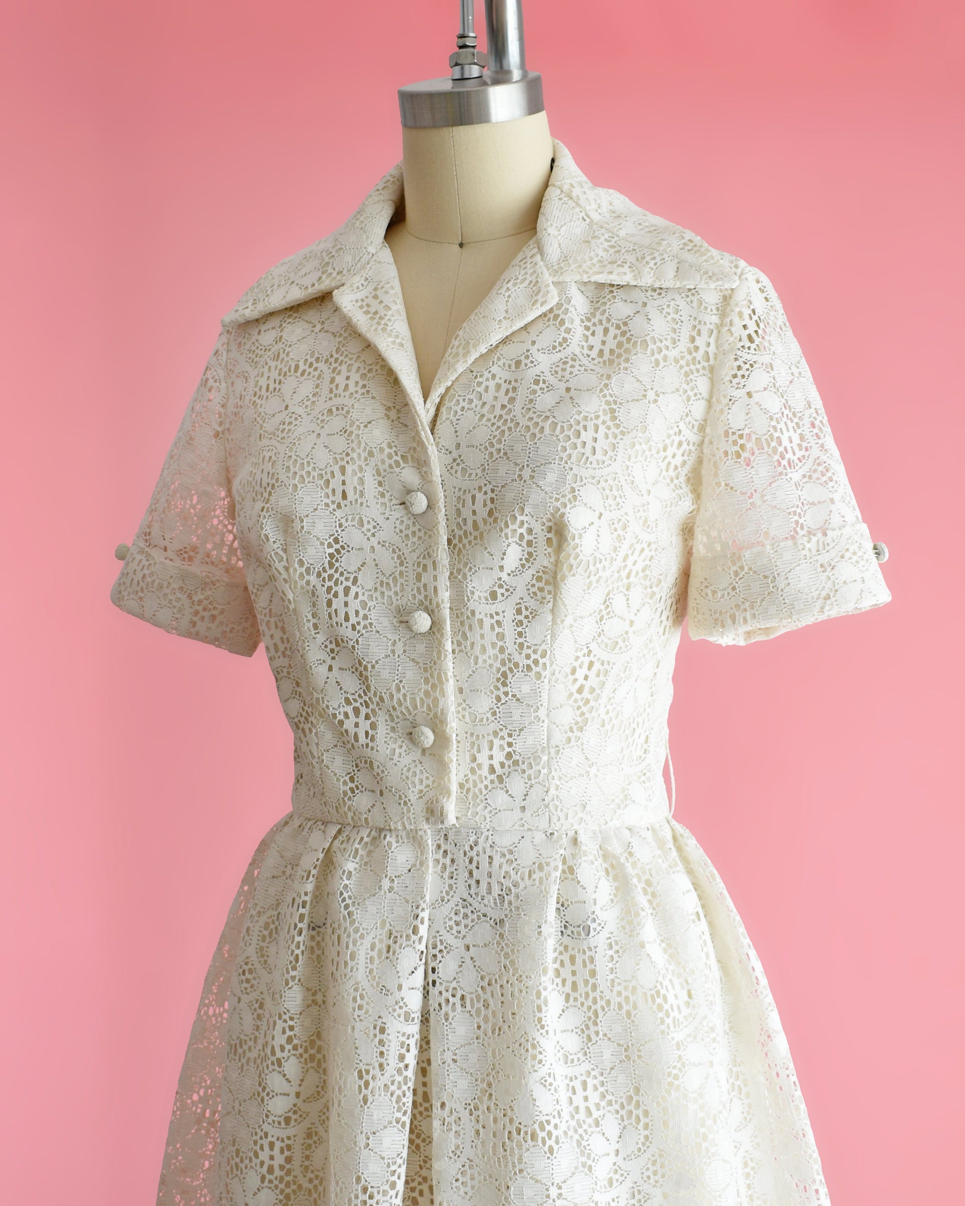 Side front view of a vintage cream floral lace dress with collared neckline, short sleeves, and button front on a dress form set on a pink background