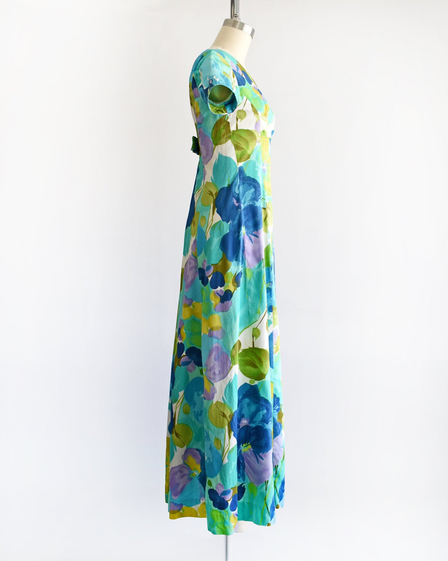 Side view of a vintage Hawaiian maxi dress on a dress form. The dress is covered with blue and green flowers that looks like a watercolor print. The dress has short sleeves, an empire waist, and a long skirt.