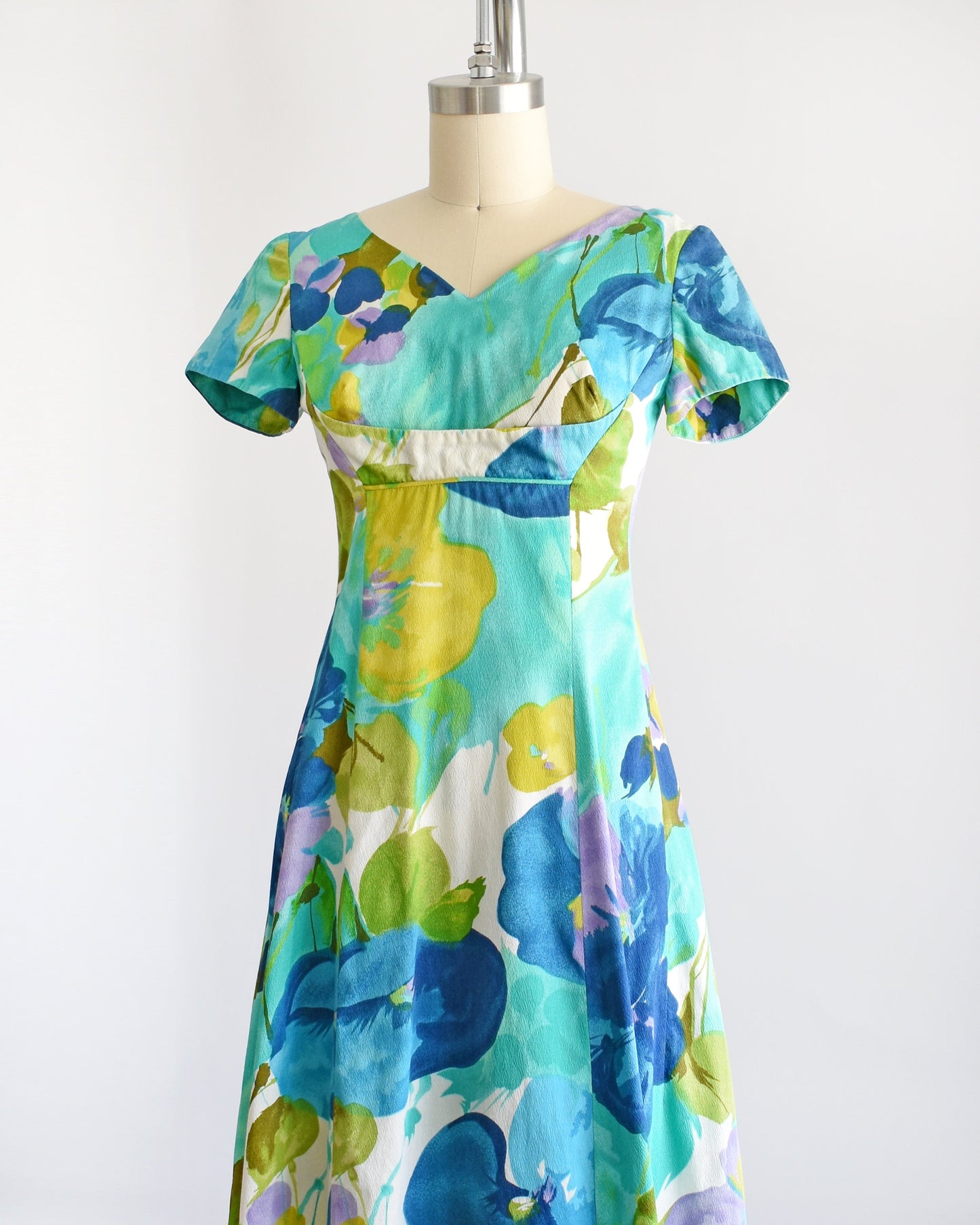 Side view of a vintage Hawaiian maxi dress on a dress form. The dress is covered with blue and green flowers that looks like a watercolor print. The dress has short sleeves, an empire waist, and a long skirt.