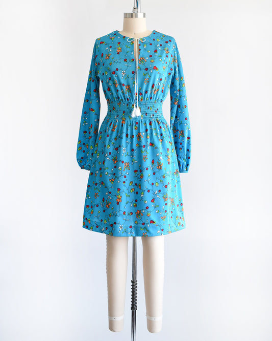 A vintage blue dress with a vine floral print with tassel neckline, smocked waist, long sleeves, and a skirt that hits above the knees. The dress is modeled on a dress form.