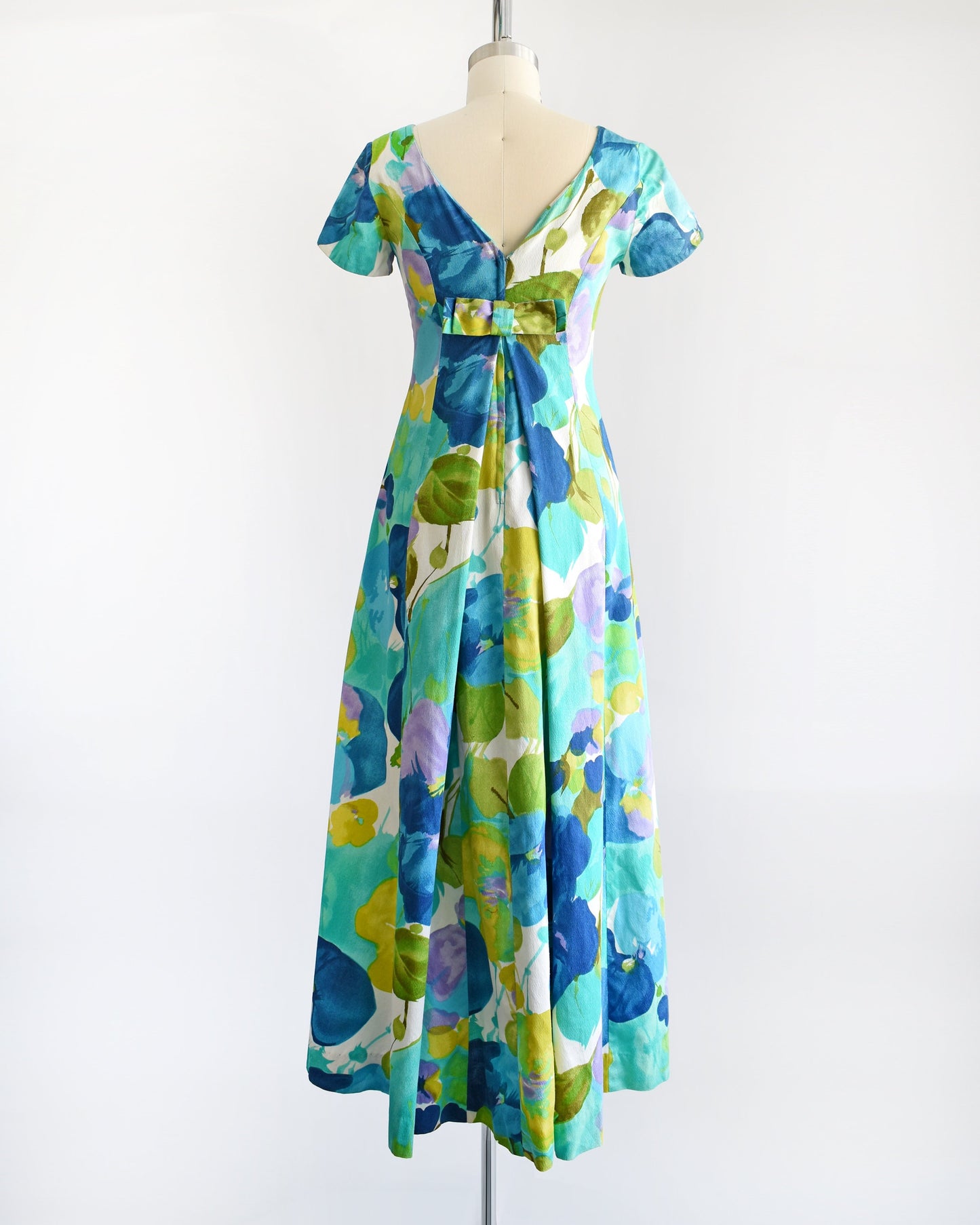 Back view of a vintage Hawaiian maxi dress on a dress form. The dress is covered with blue and green flowers that looks like a watercolor print. The dress has short sleeves, an empire waist, and a long skirt. The back has a bow above the waist.